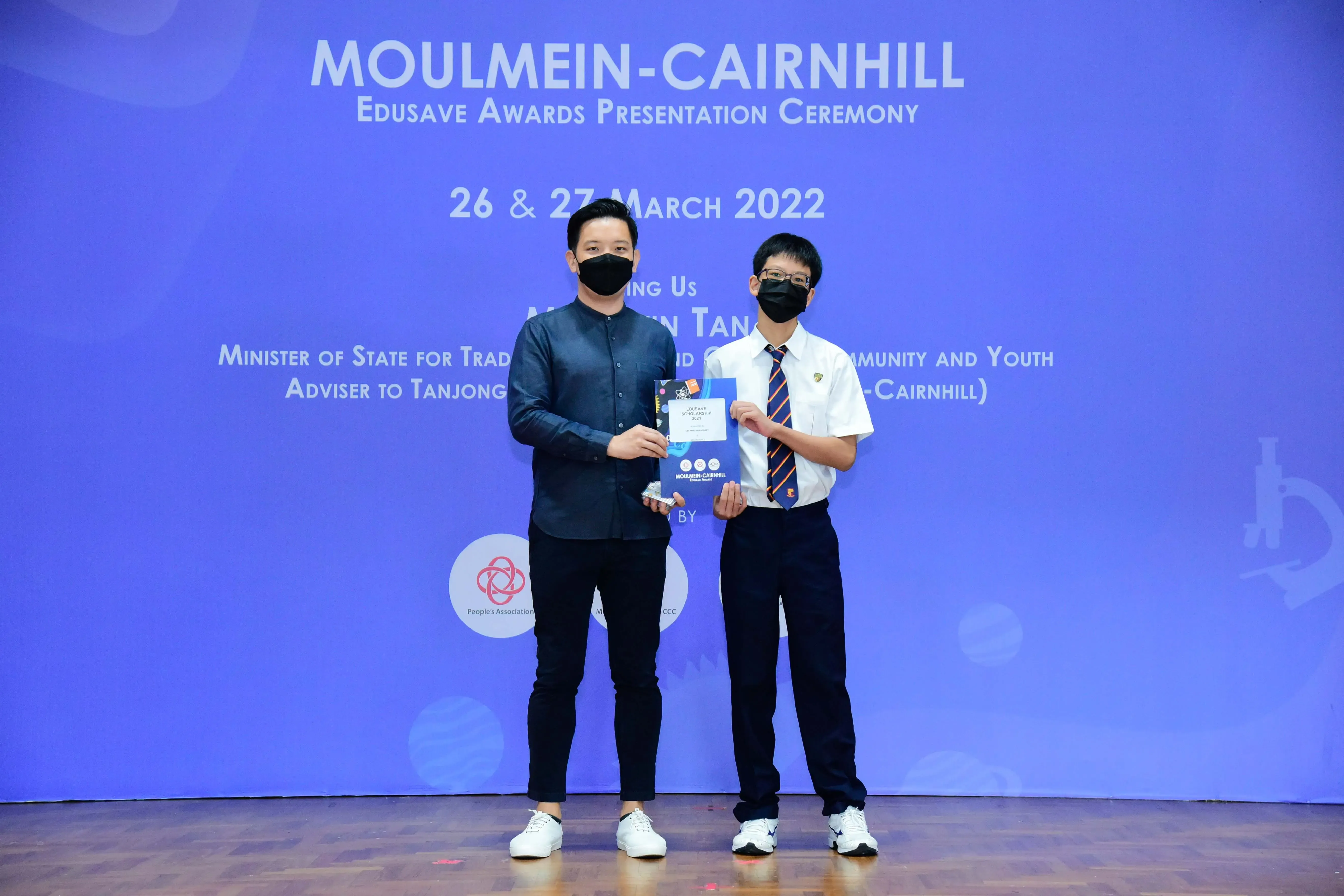Two masked individuals hold an award at the Moulmein-Cairnhill Edusave Awards Presentation Ceremony, March 2022.
