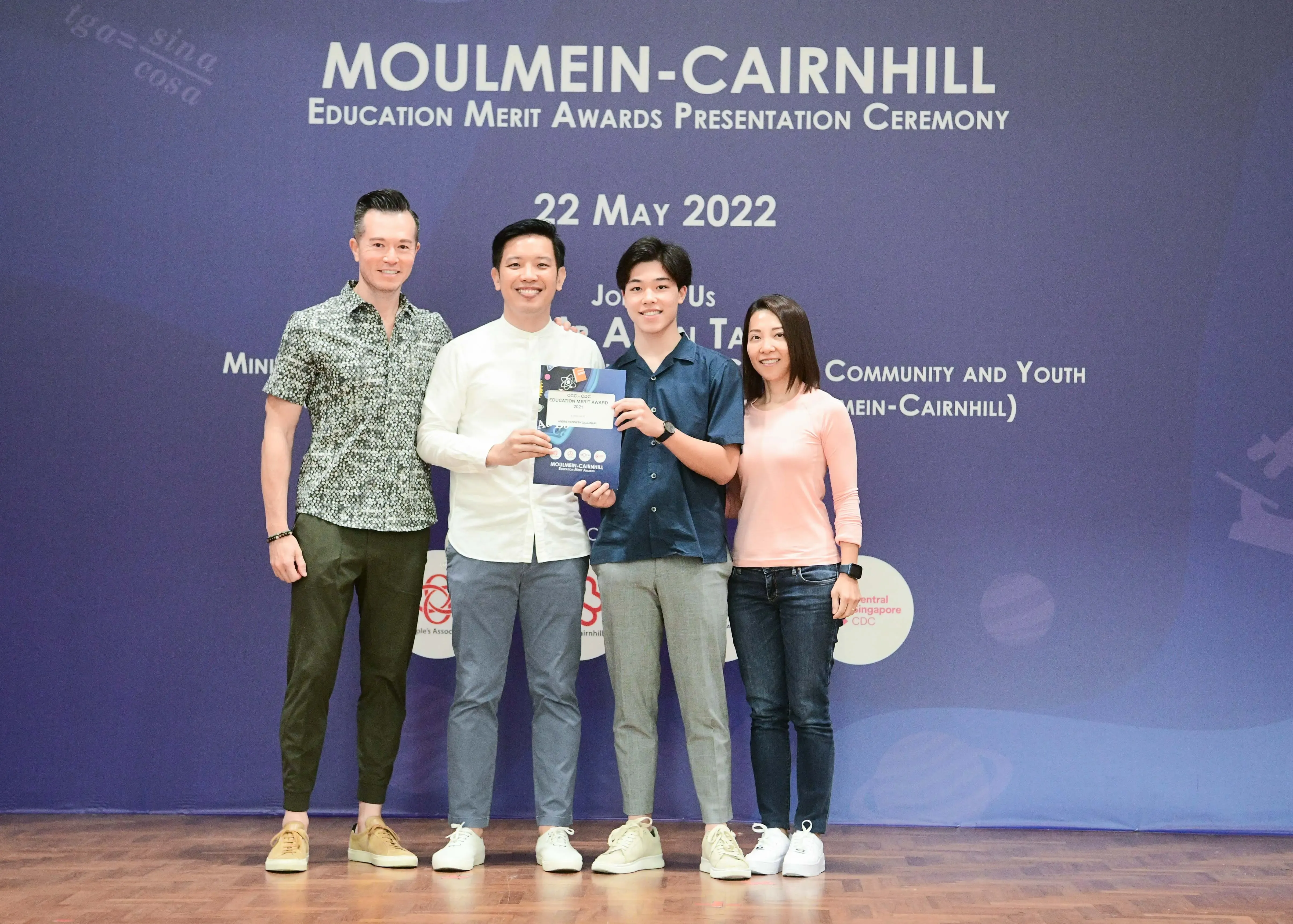 Four people pose smiling before a Moulmein-Cairnhill Education Merit Awards banner dated 22 May 2022, holding a certificate.