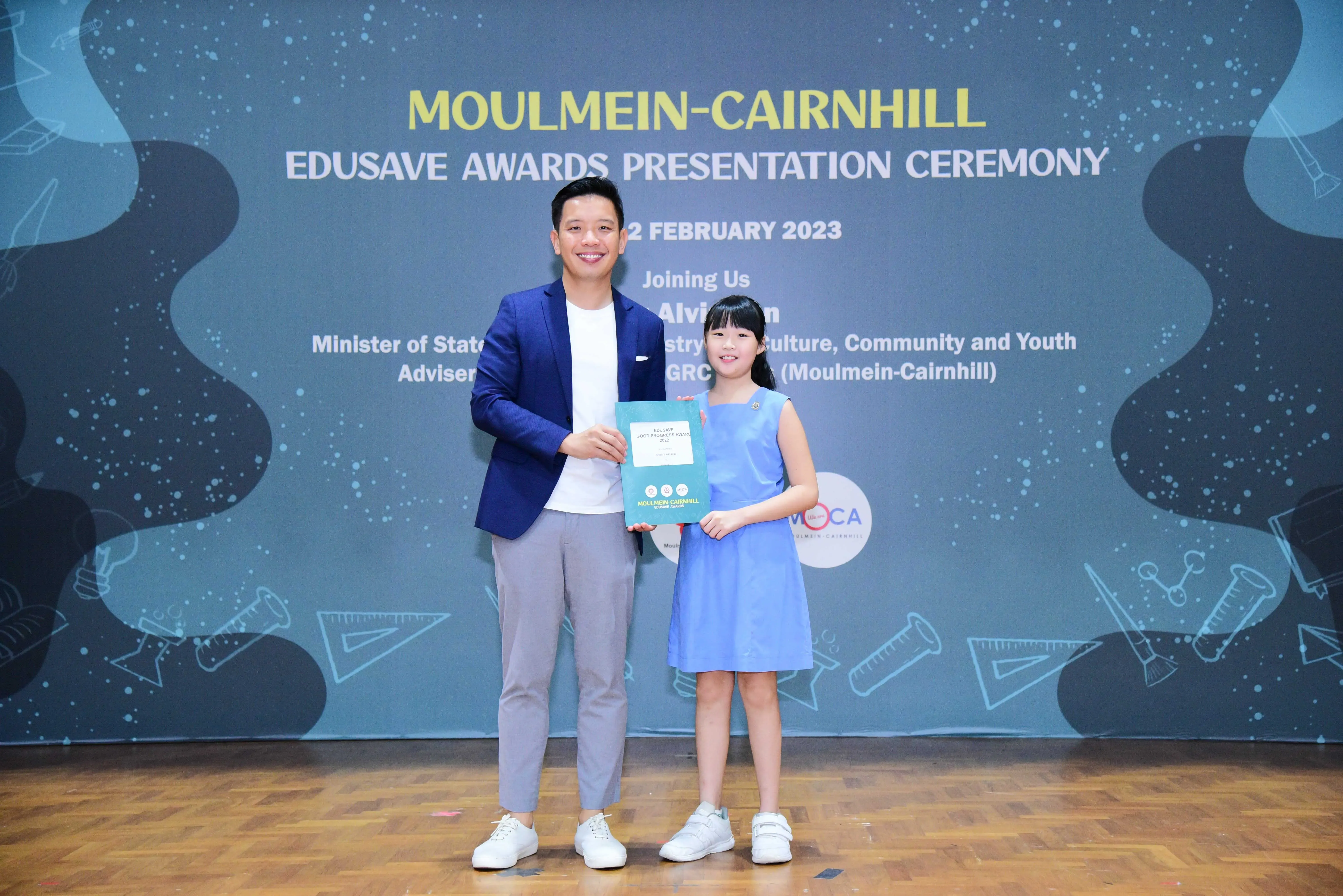 A man in a blue blazer and a young girl in a blue dress hold a certificate at the Moulmein-Cairnhill Edusave Awards 2023.