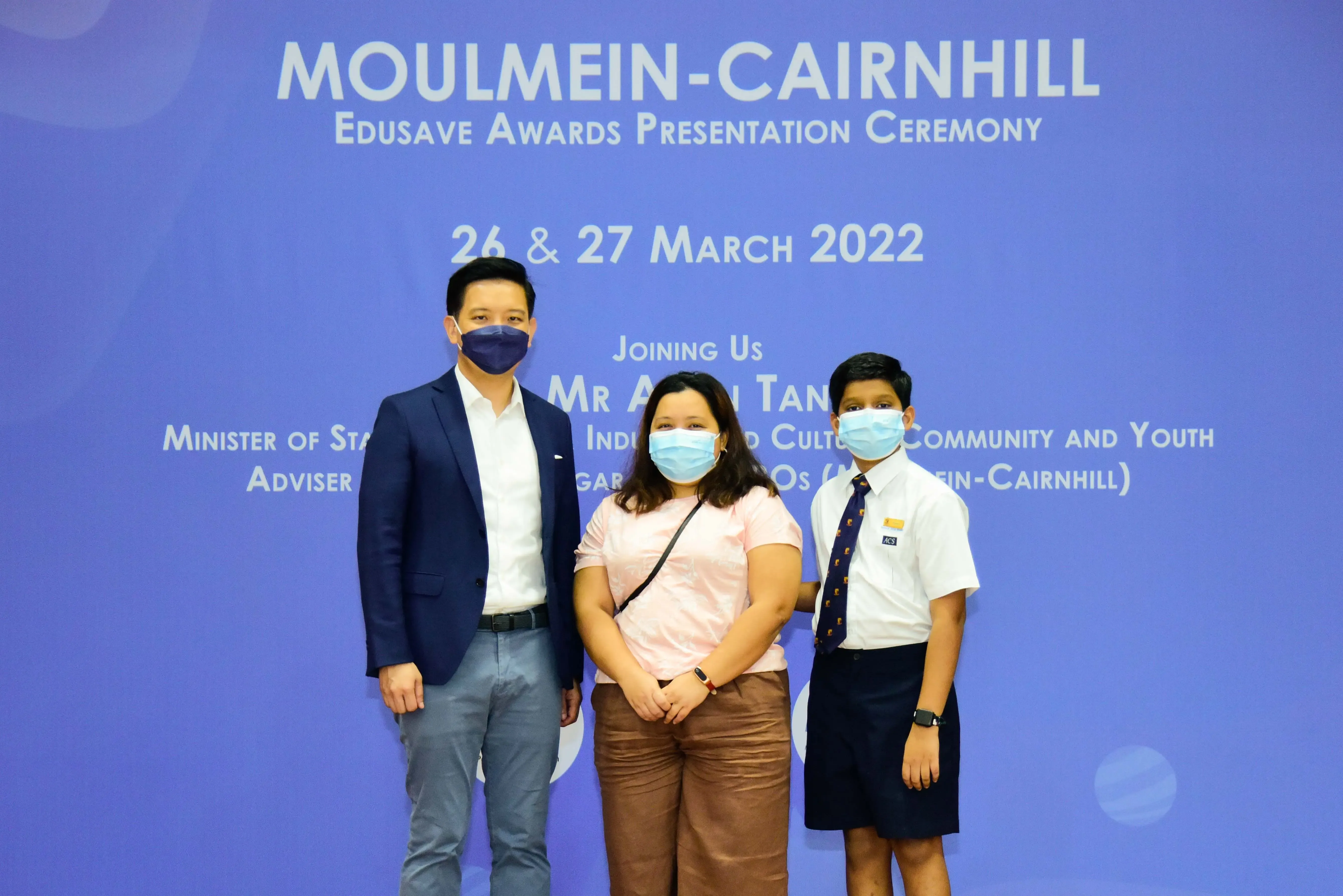 Three masked people pose before a blue Moulmein-Cairnhill Edusave Awards Presentation Ceremony banner dated March 2022.