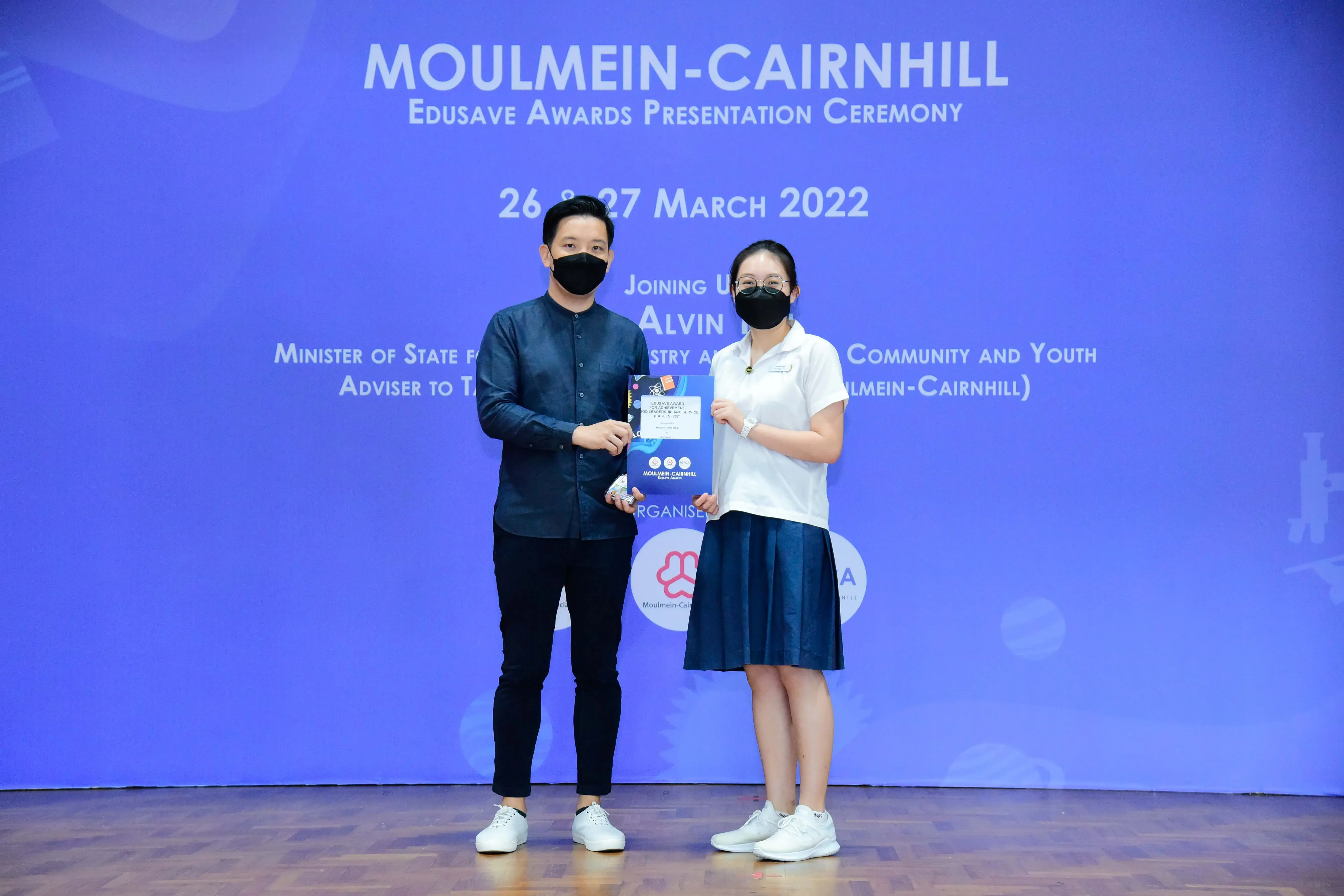 Two masked people hold a certificate at the Moulmein-Cairnhill Edusave Awards Presentation Ceremony, March 2022.