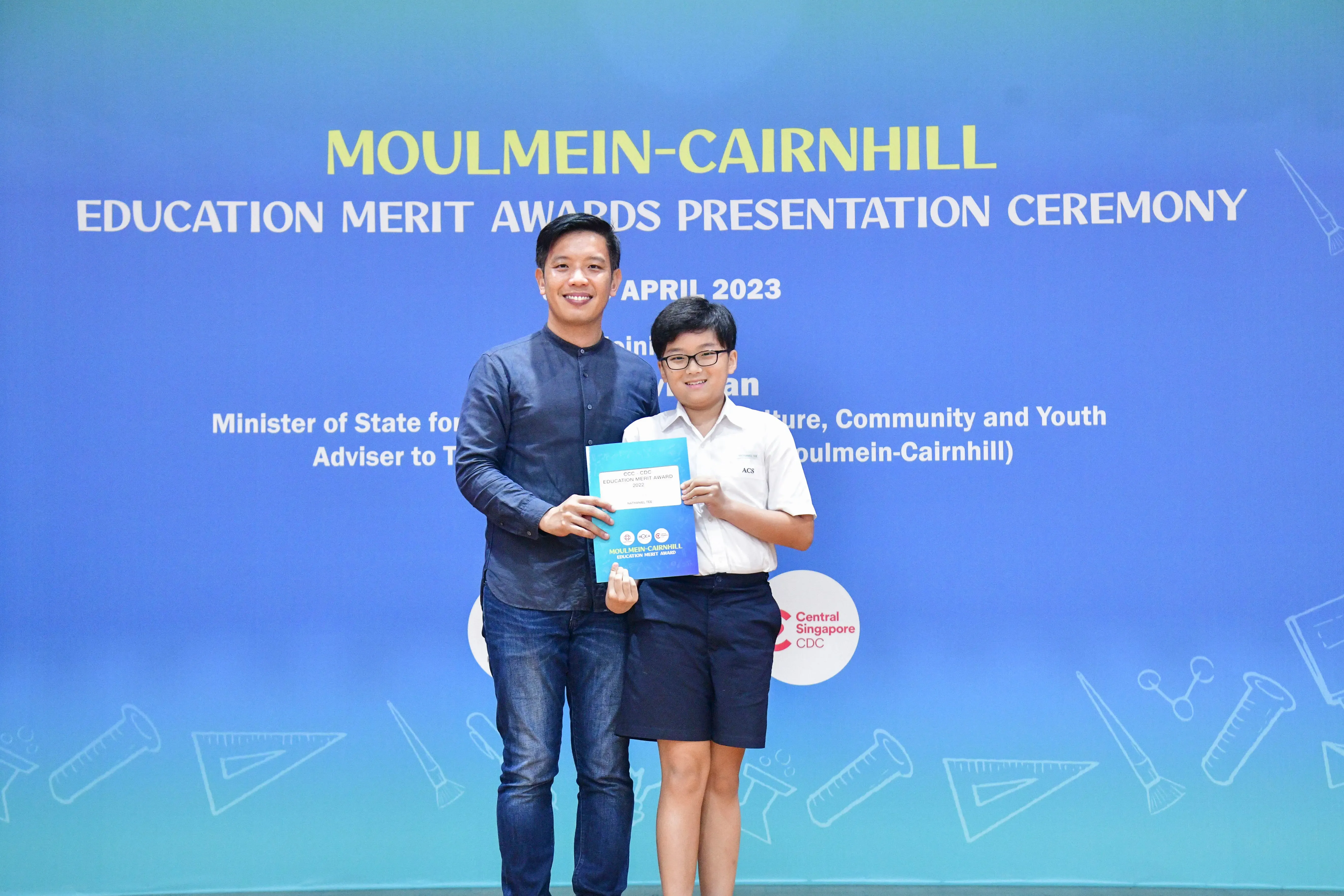 An adult and a young student hold an award certificate at the Moulmein-Cairnhill Education Merit Awards Ceremony, April 2023.