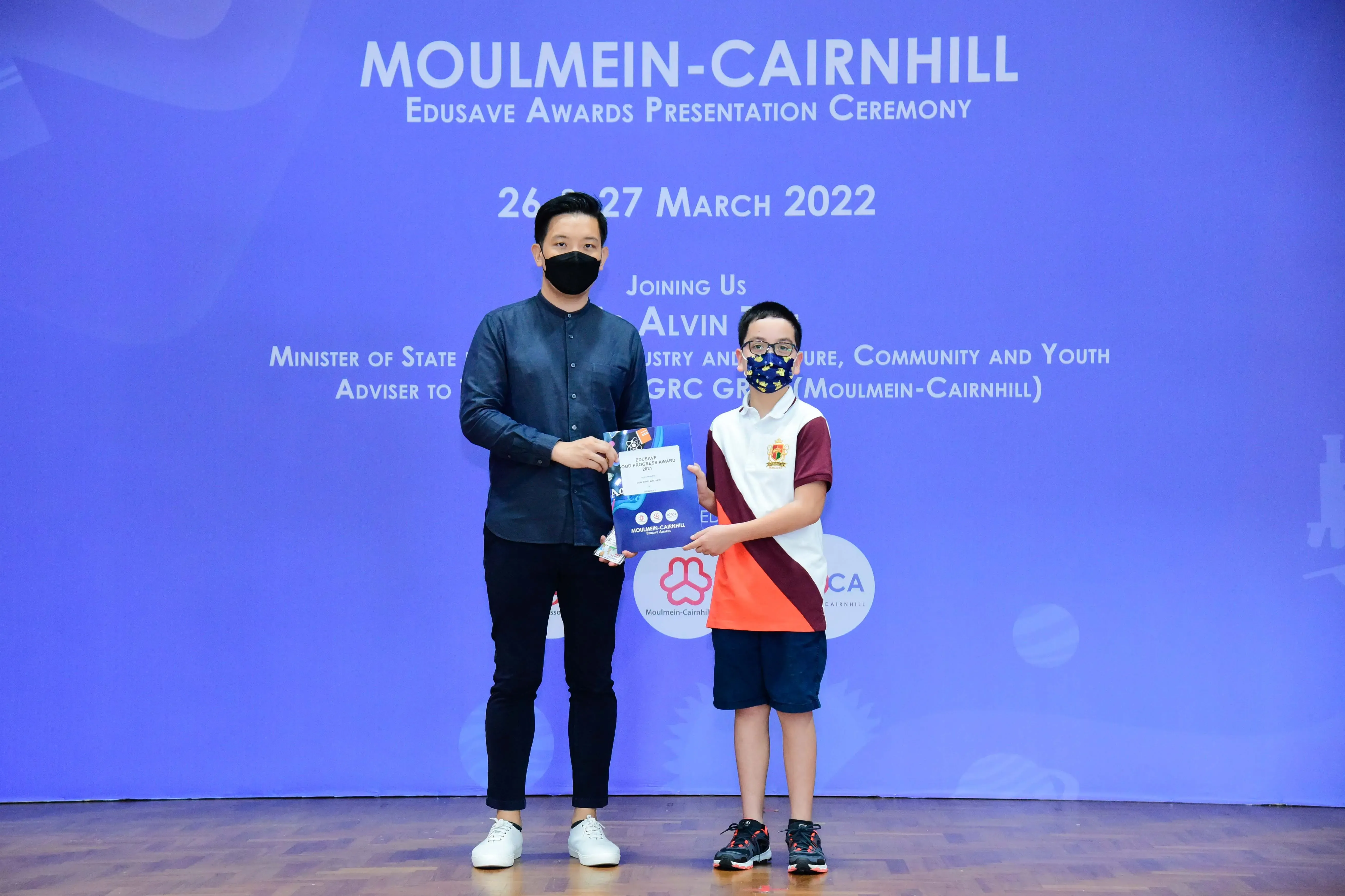 An adult and a masked child hold a certificate at the Moulmein-Cairnhill Edusave Awards Ceremony, March 2022.