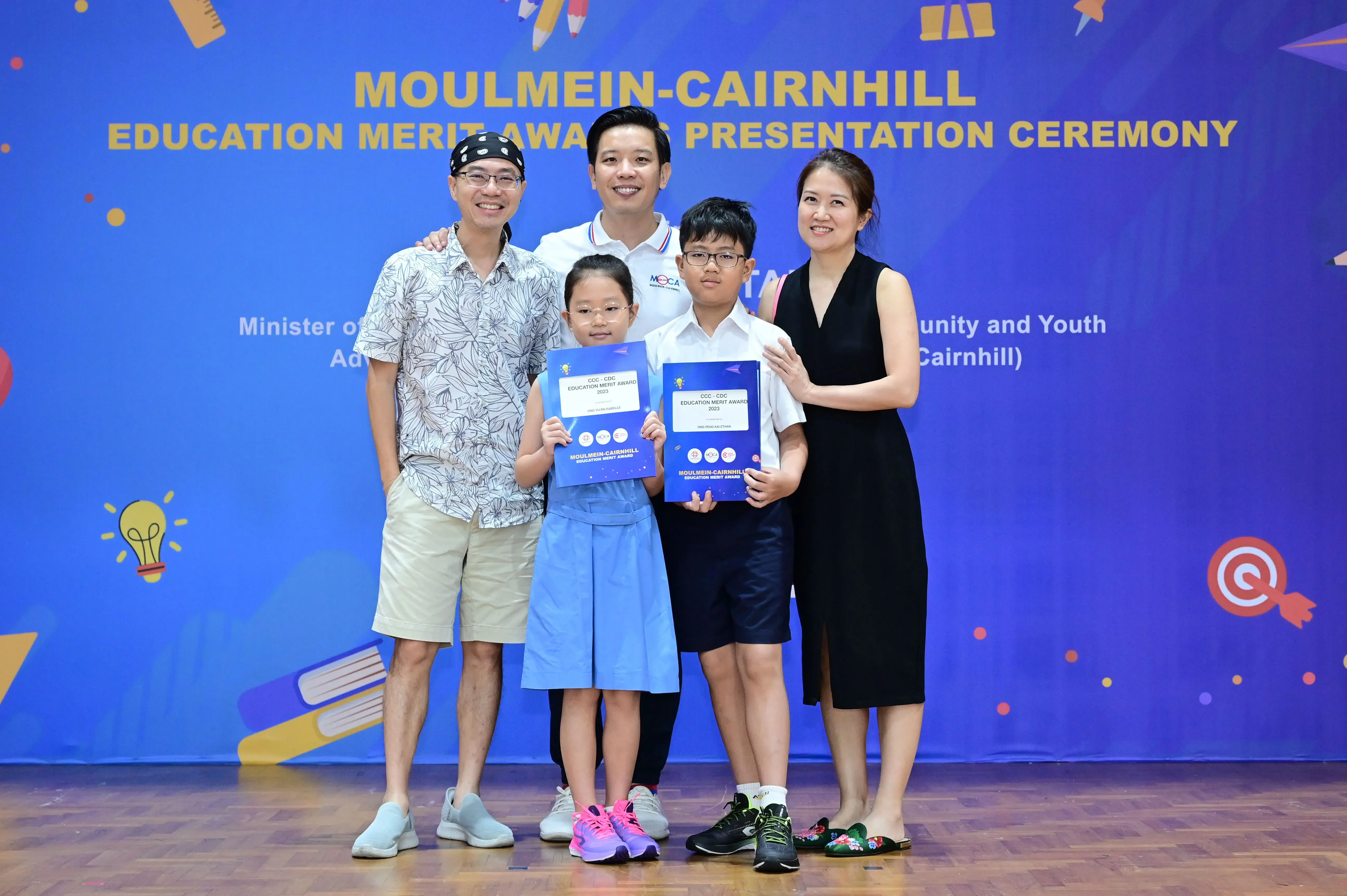 Five people pose at the Moulmein-Cairnhill Education Merit Awards ceremony, two children holding award folders.