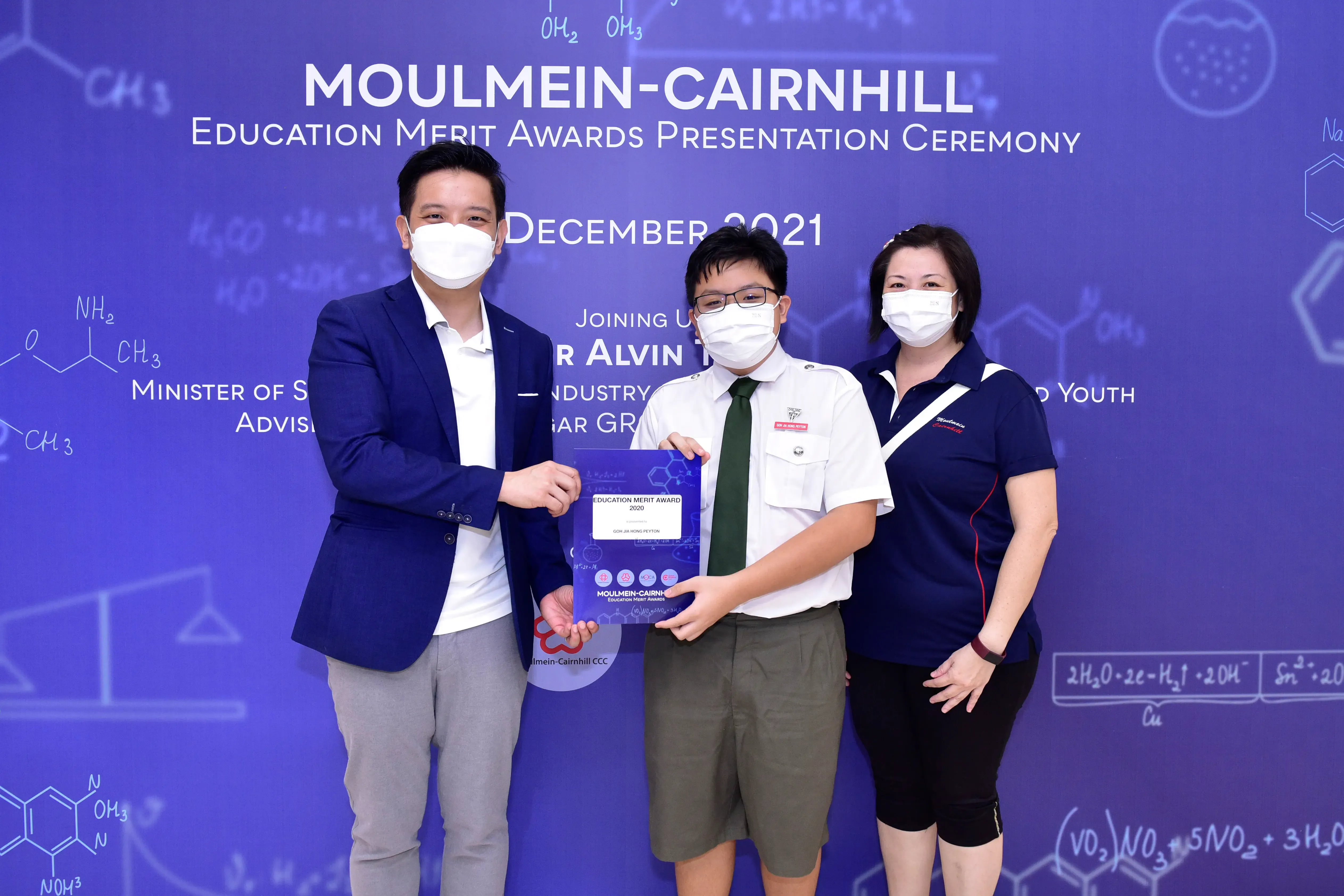 Three masked people pose at the Moulmein-Cairnhill Education Merit Awards Ceremony, December 2021, holding a certificate.