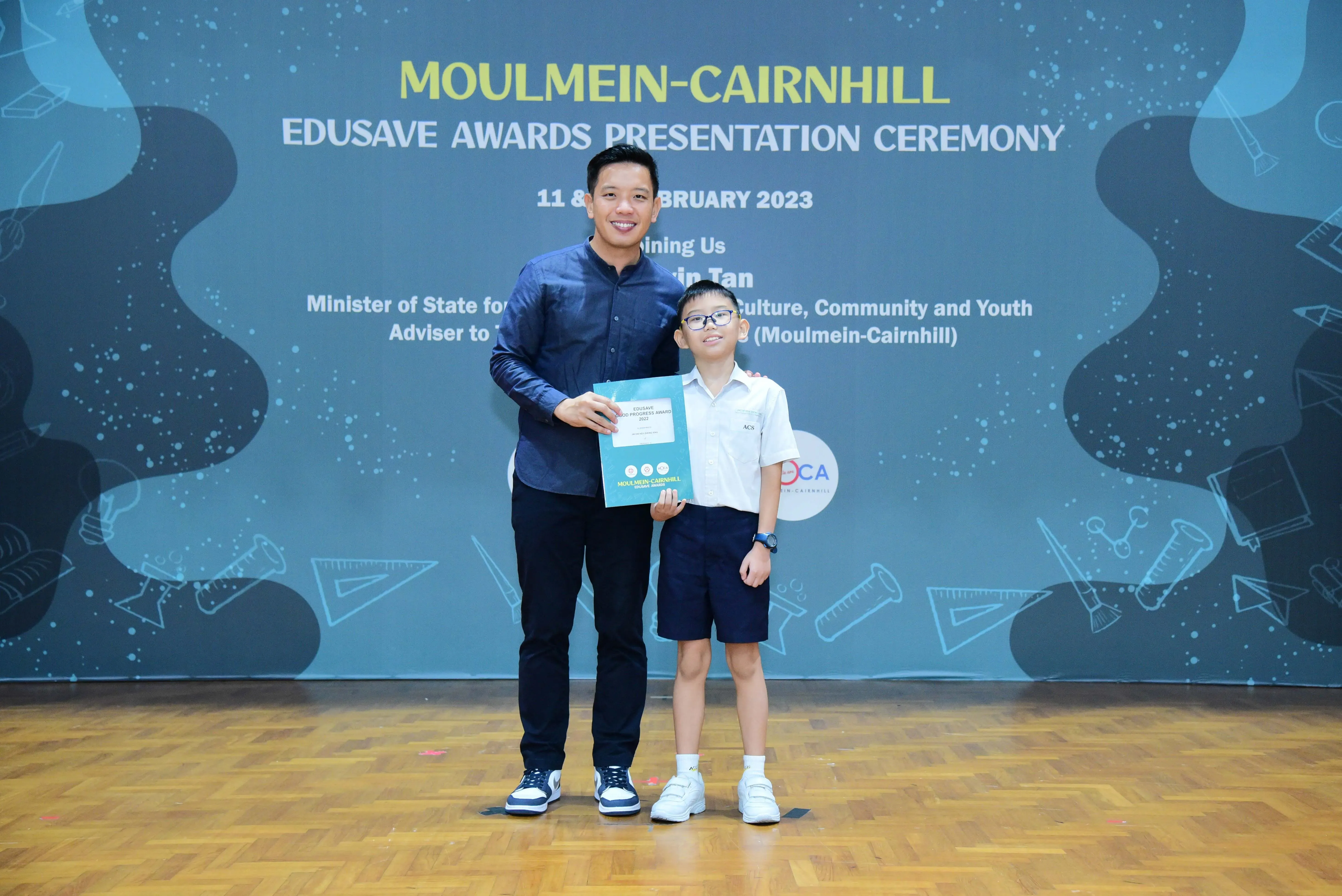 An adult and a young boy in school uniform hold a certificate at the Moulmein-Cairnhill Edusave Awards Ceremony 2023.
