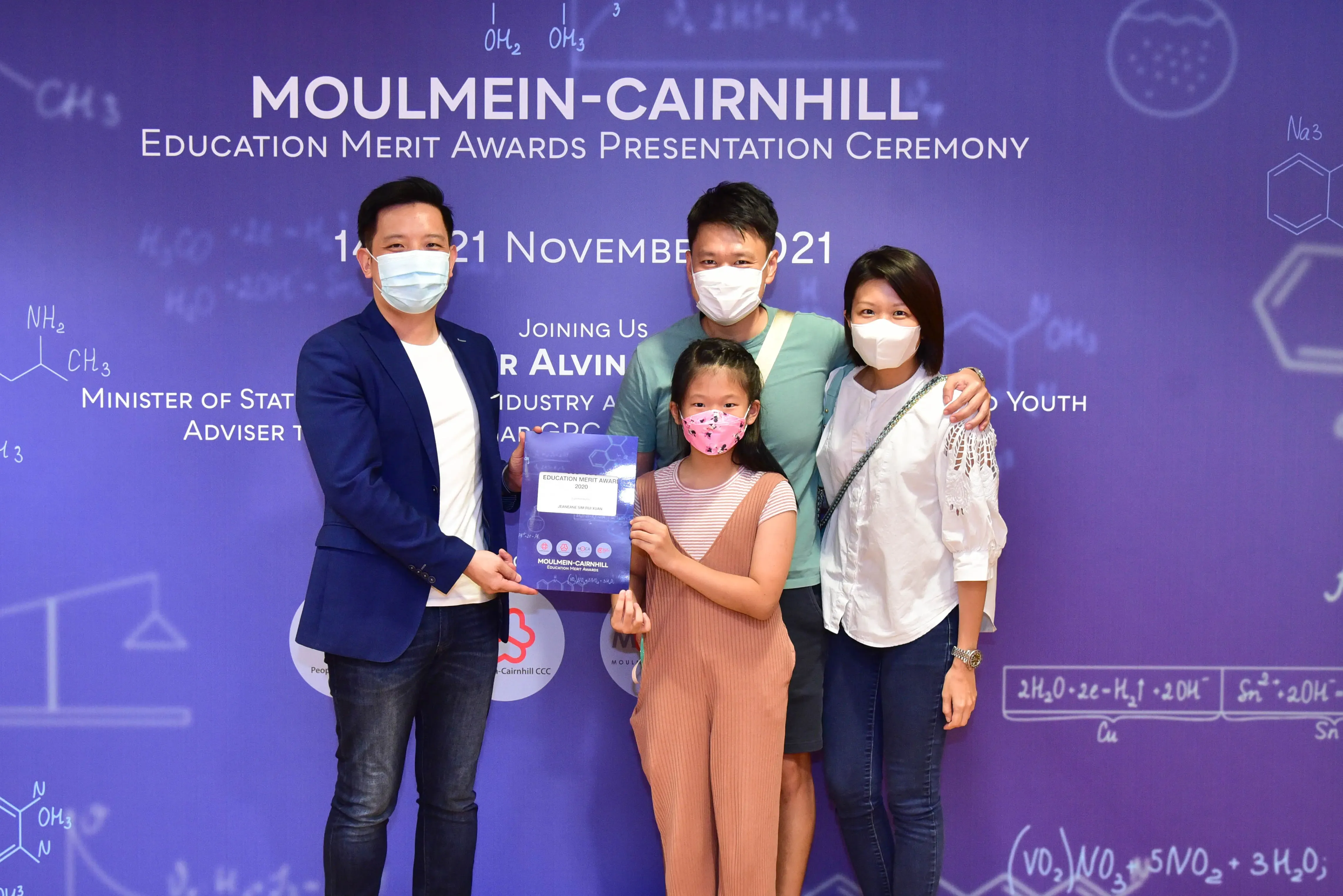 Four masked people pose at the Moulmein-Cairnhill Education Merit Awards Ceremony, with a girl holding an award certificate.