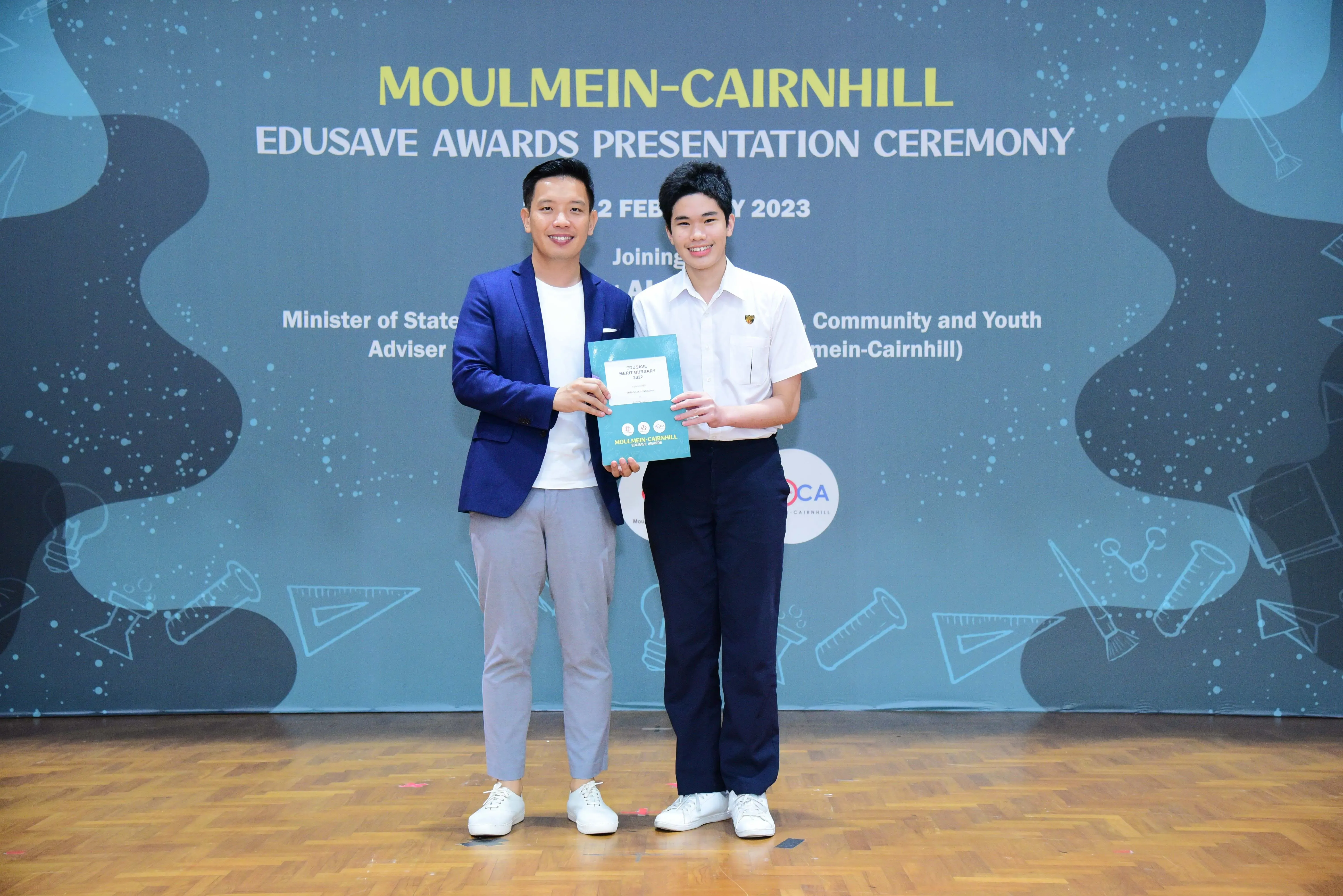 Two men smile holding an award booklet at the Moulmein-Cairnhill Edusave Awards Presentation Ceremony 2023.