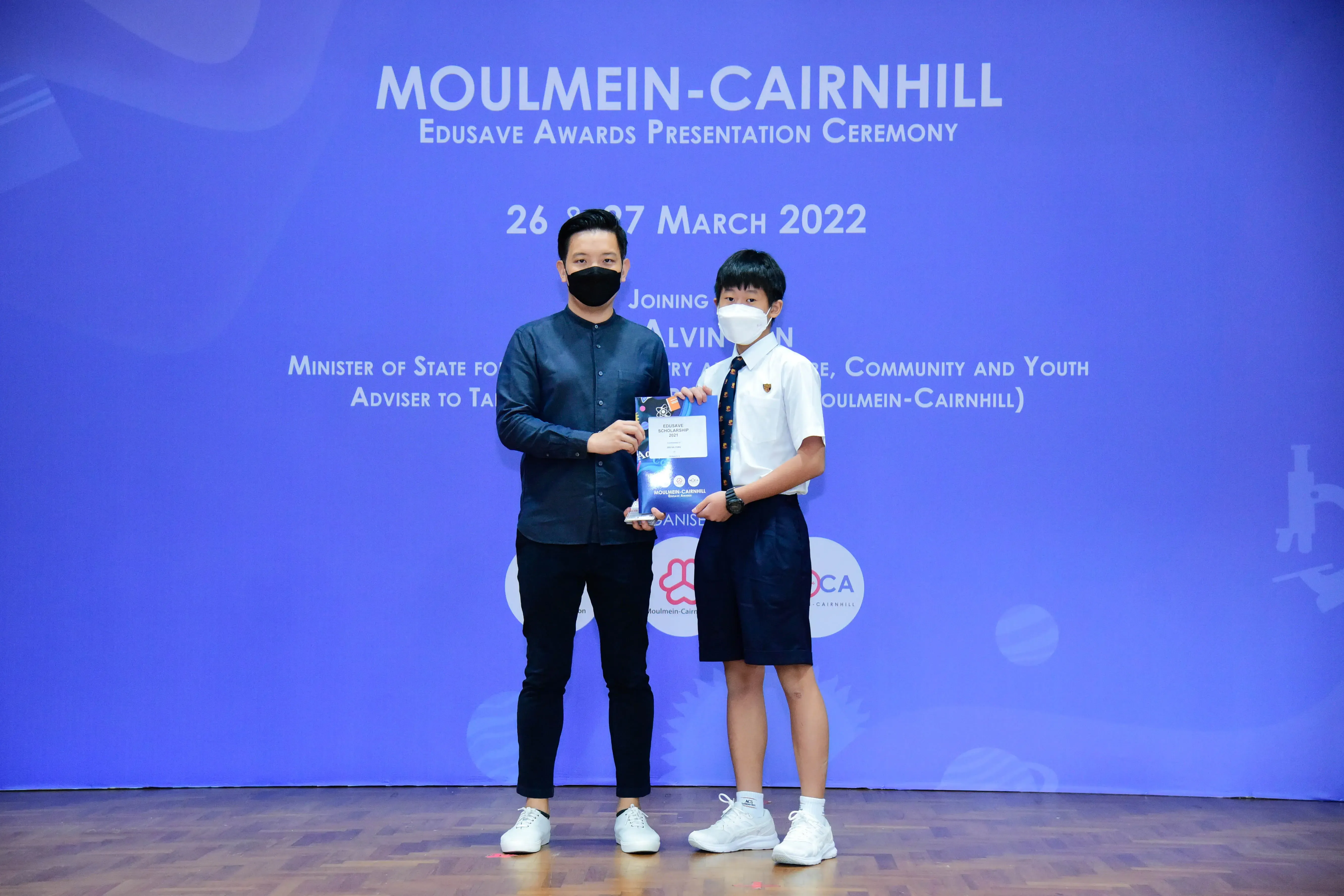 Two masked individuals hold an award at the Moulmein-Cairnhill Edusave Awards Presentation Ceremony, March 2022.
