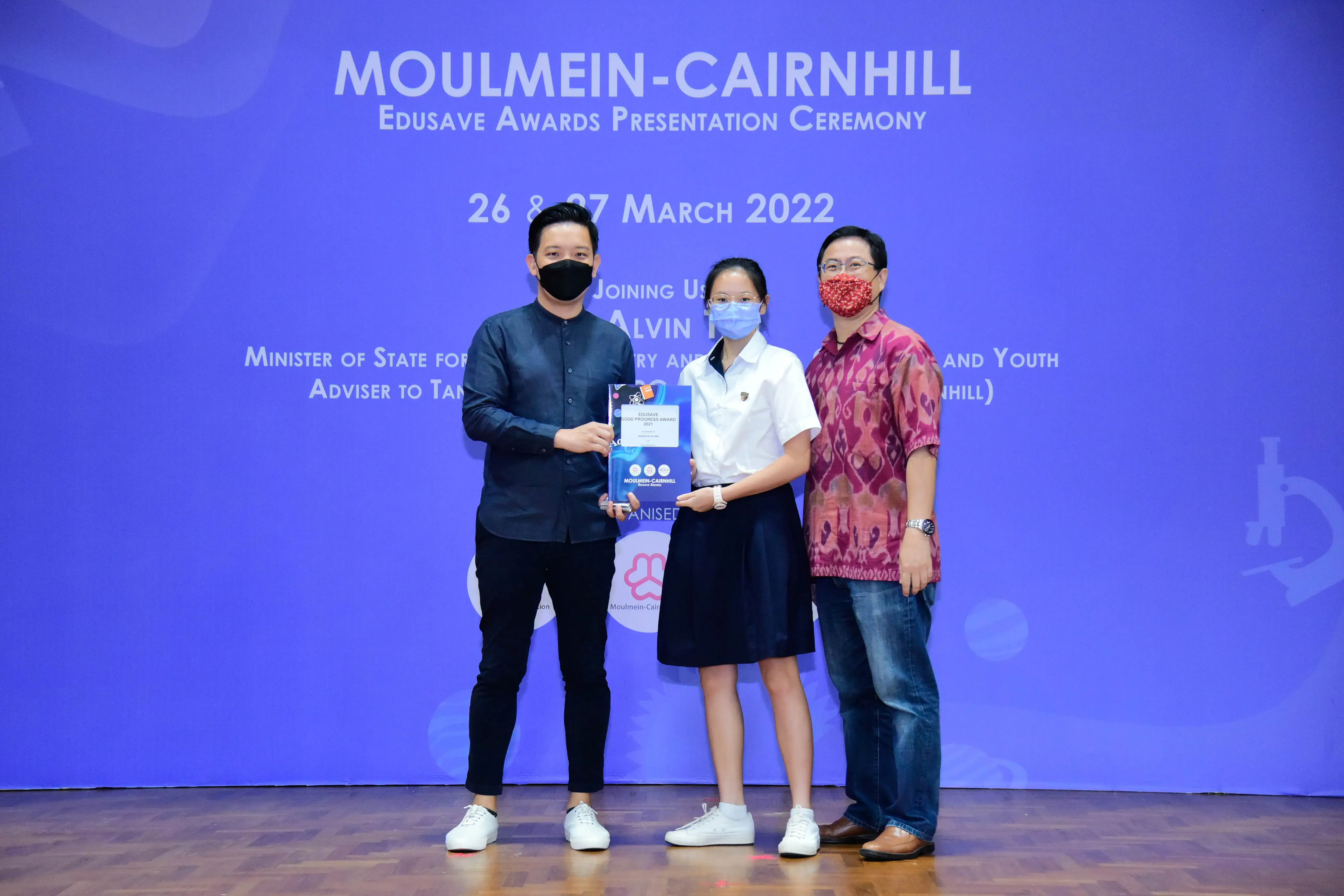 Three masked people stand on a stage holding an award booklet at the Moulmein-Cairnhill Edusave Awards Ceremony, March 2022.