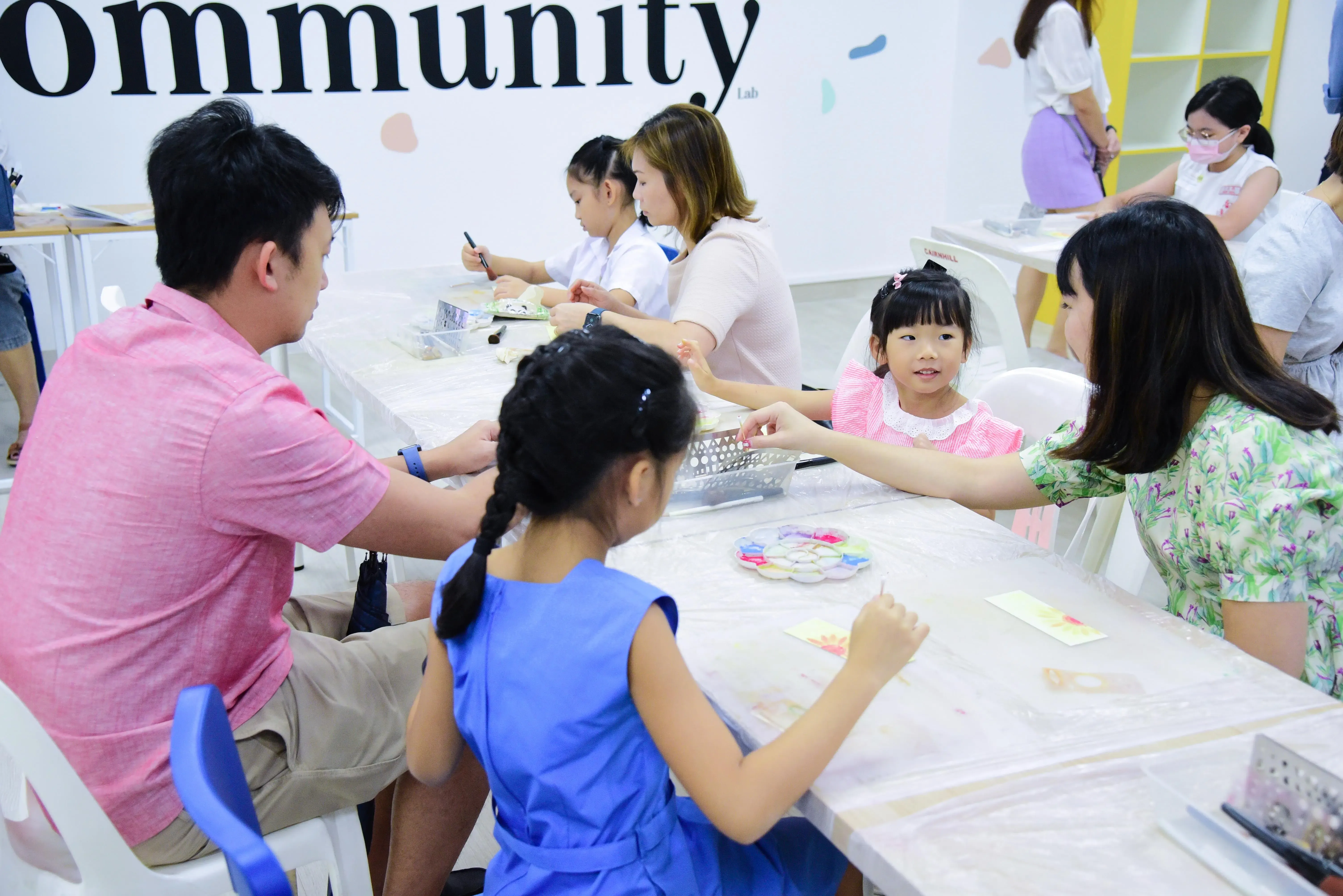 Adults and children sit at tables doing an art craft activity in a bright room with "community Lab" on the wall.