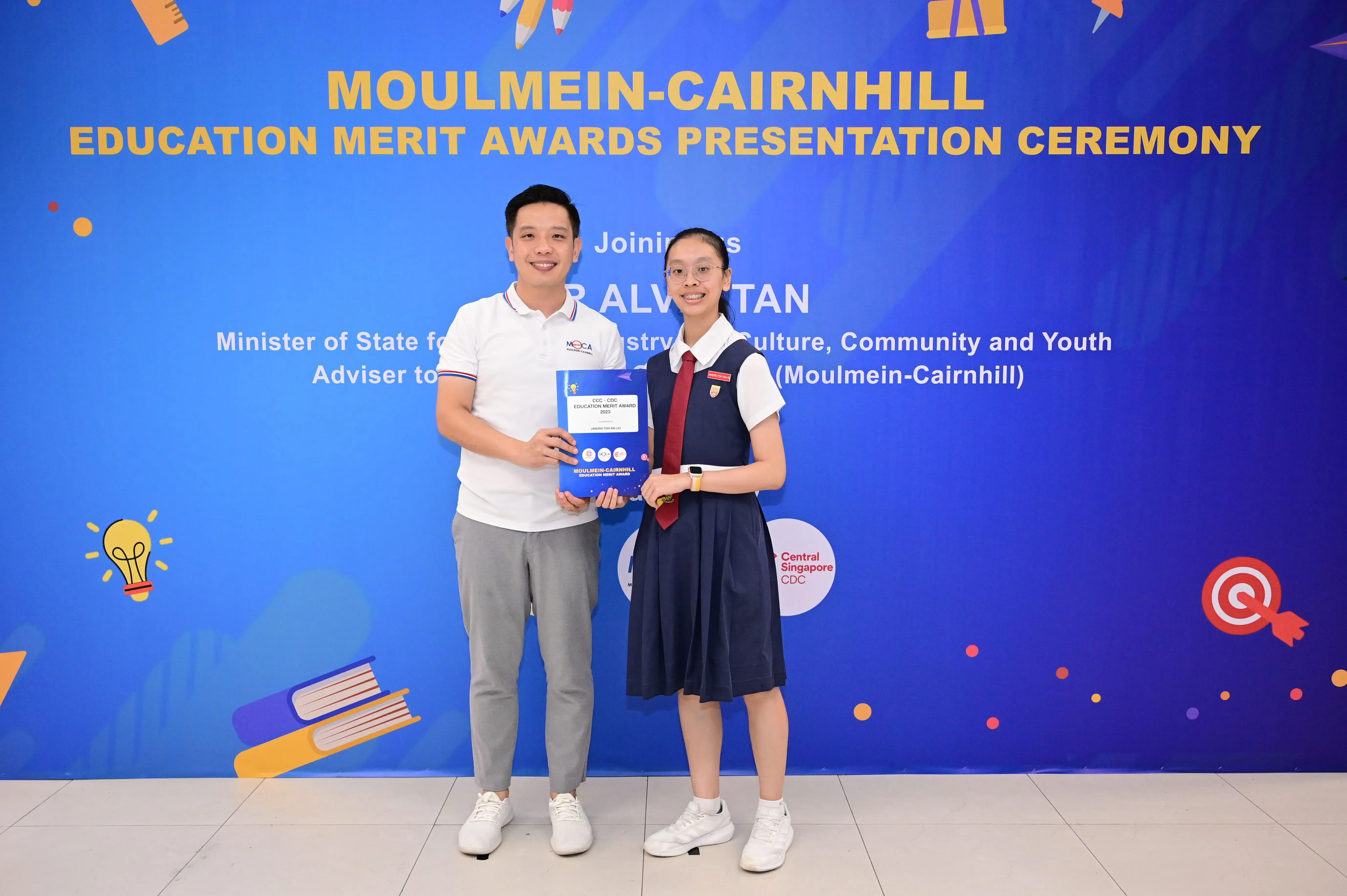 Two people hold a certificate together in front of a Moulmein-Cairnhill Education Merit Awards banner.