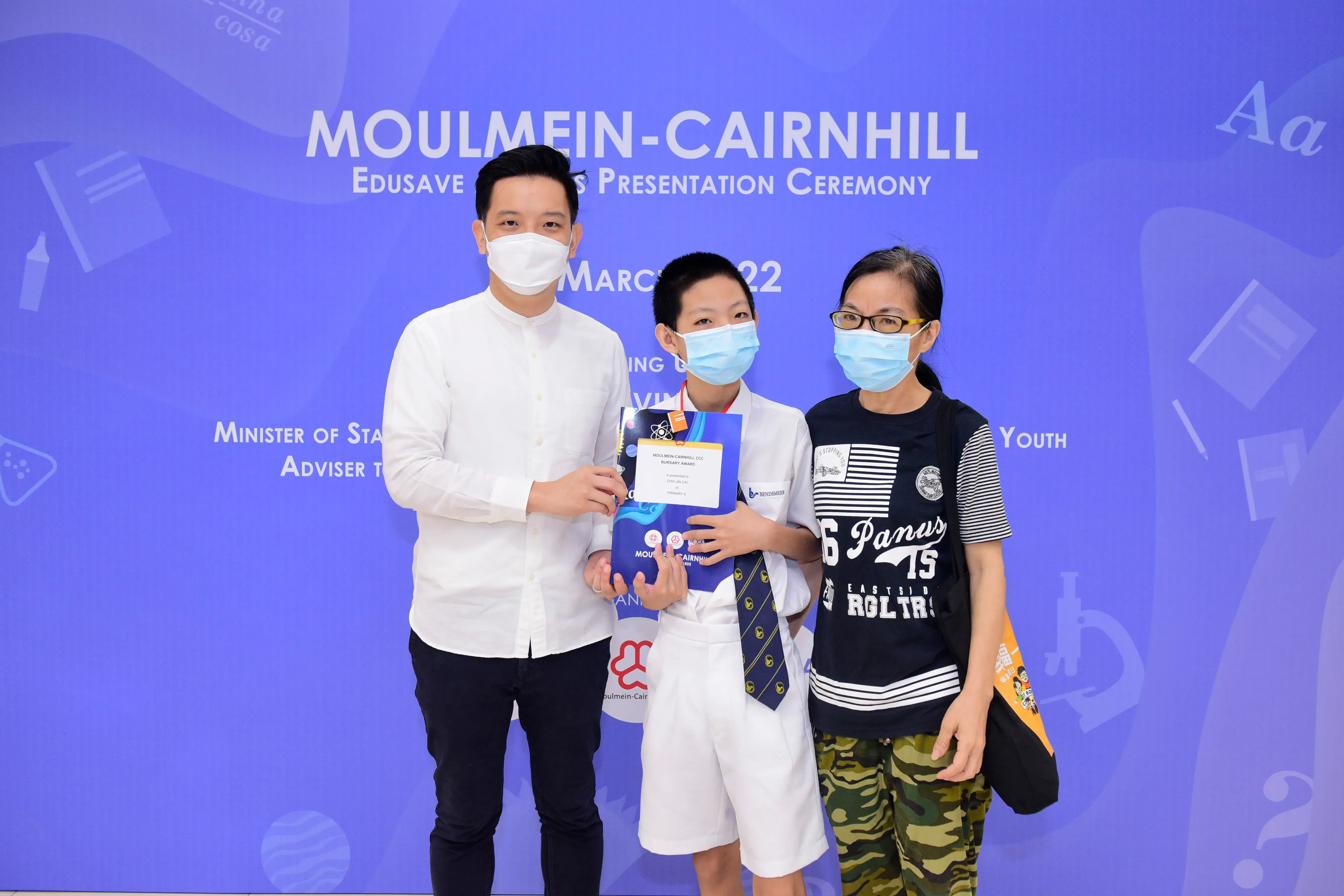 Three masked people pose at the Moulmein-Cairnhill Edusave Awards Presentation Ceremony, with a boy holding an award.