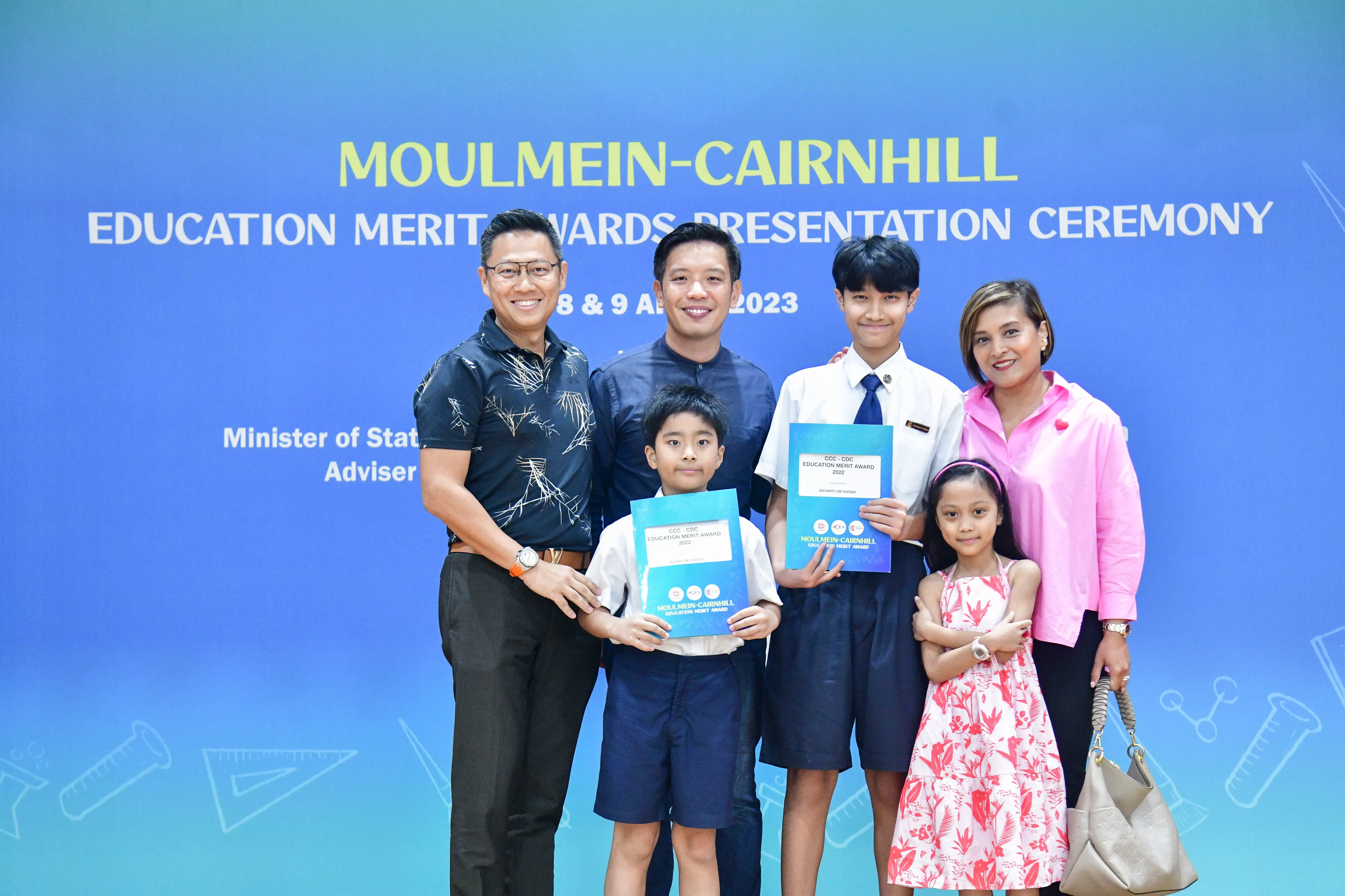 Four adults and two children pose holding award certificates at the Moulmein-Cairnhill Education Merit Awards Ceremony 2023.