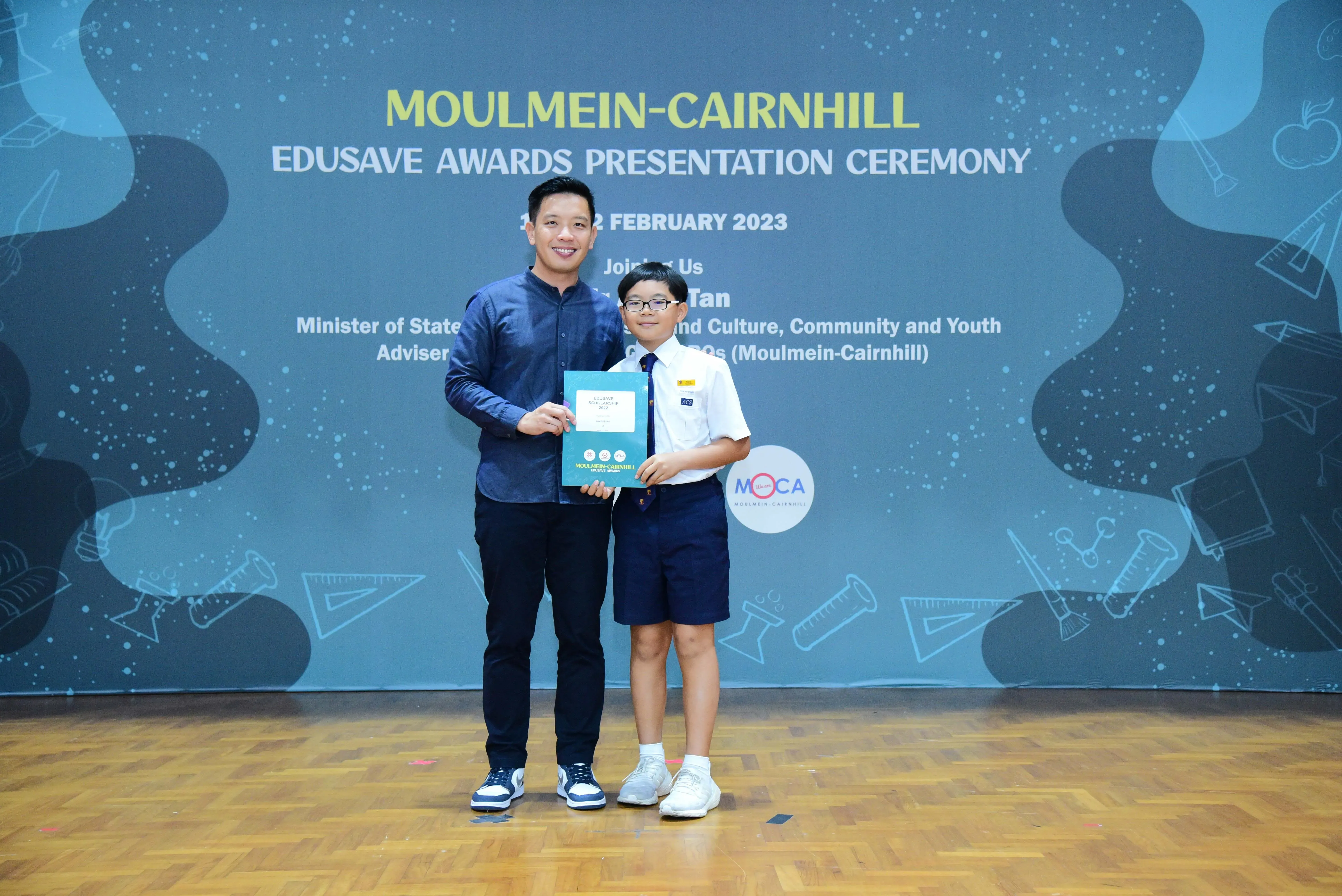 A man and a young student in school uniform hold a certificate at the Moulmein-Cairnhill Edusave Awards Ceremony 2023.