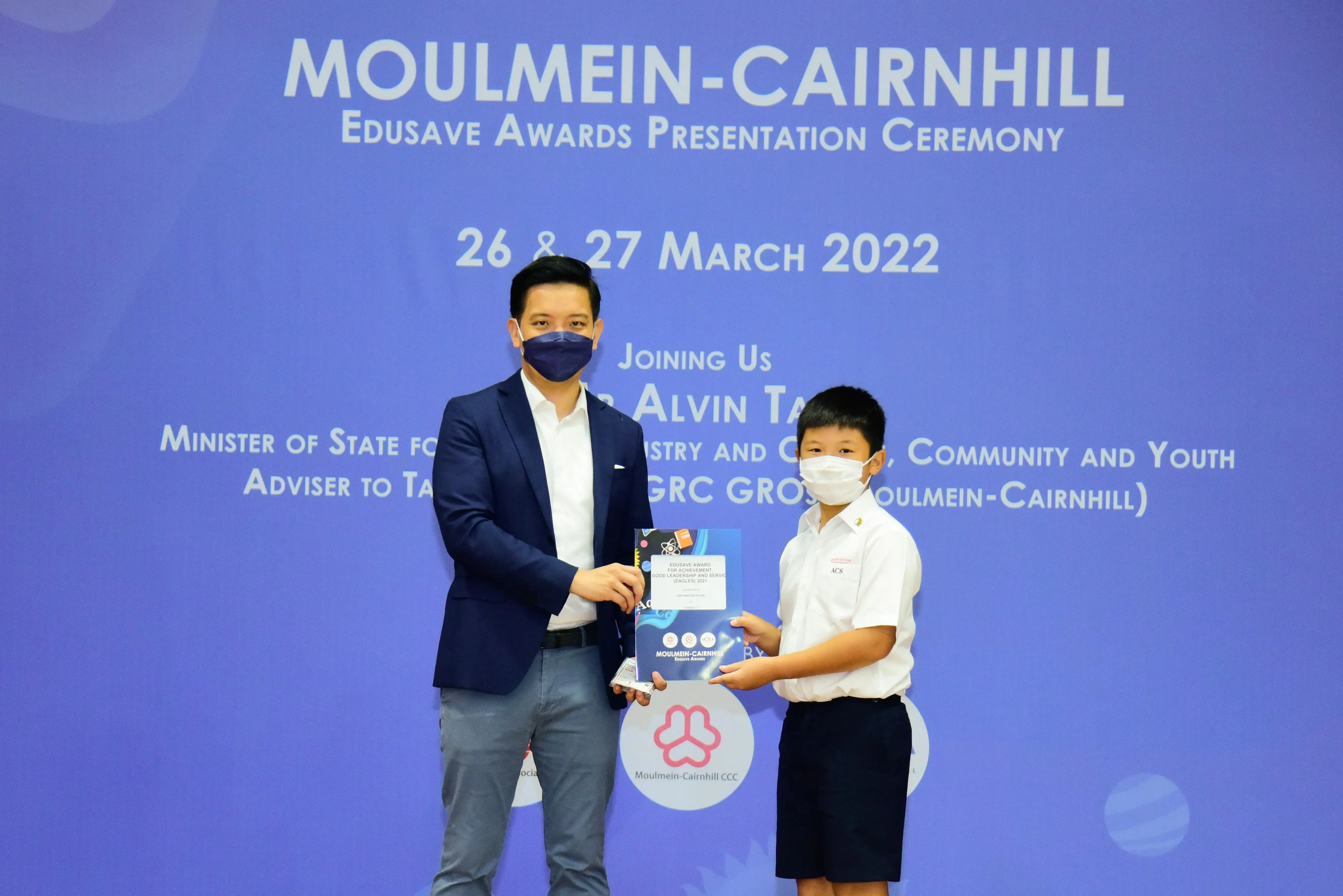 An adult and a young student, both masked, hold an award booklet at the Moulmein-Cairnhill Edusave Awards Ceremony 2022.