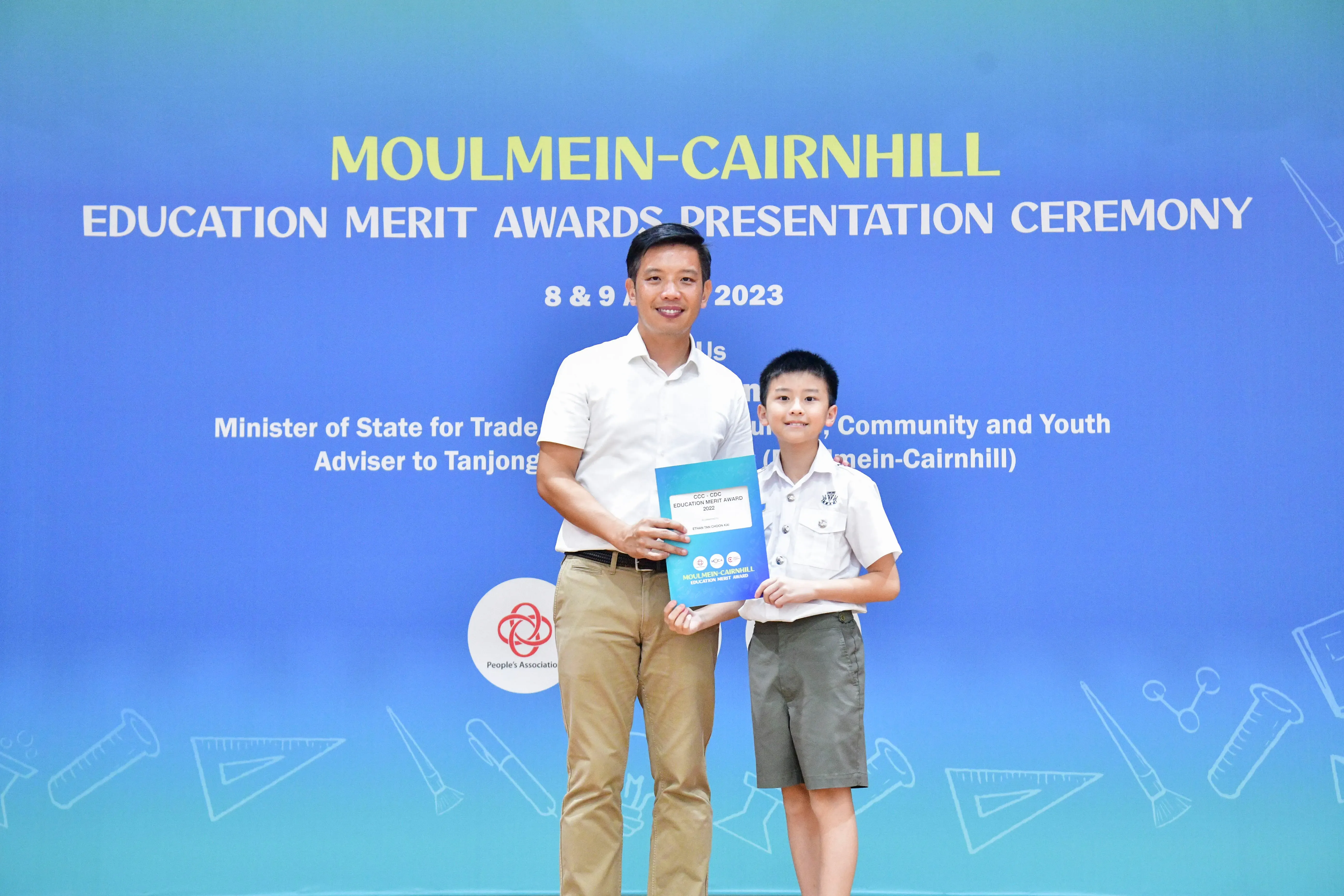 A man and a young boy hold a teal award folder at the Moulmein-Cairnhill Education Merit Awards Ceremony 2023.
