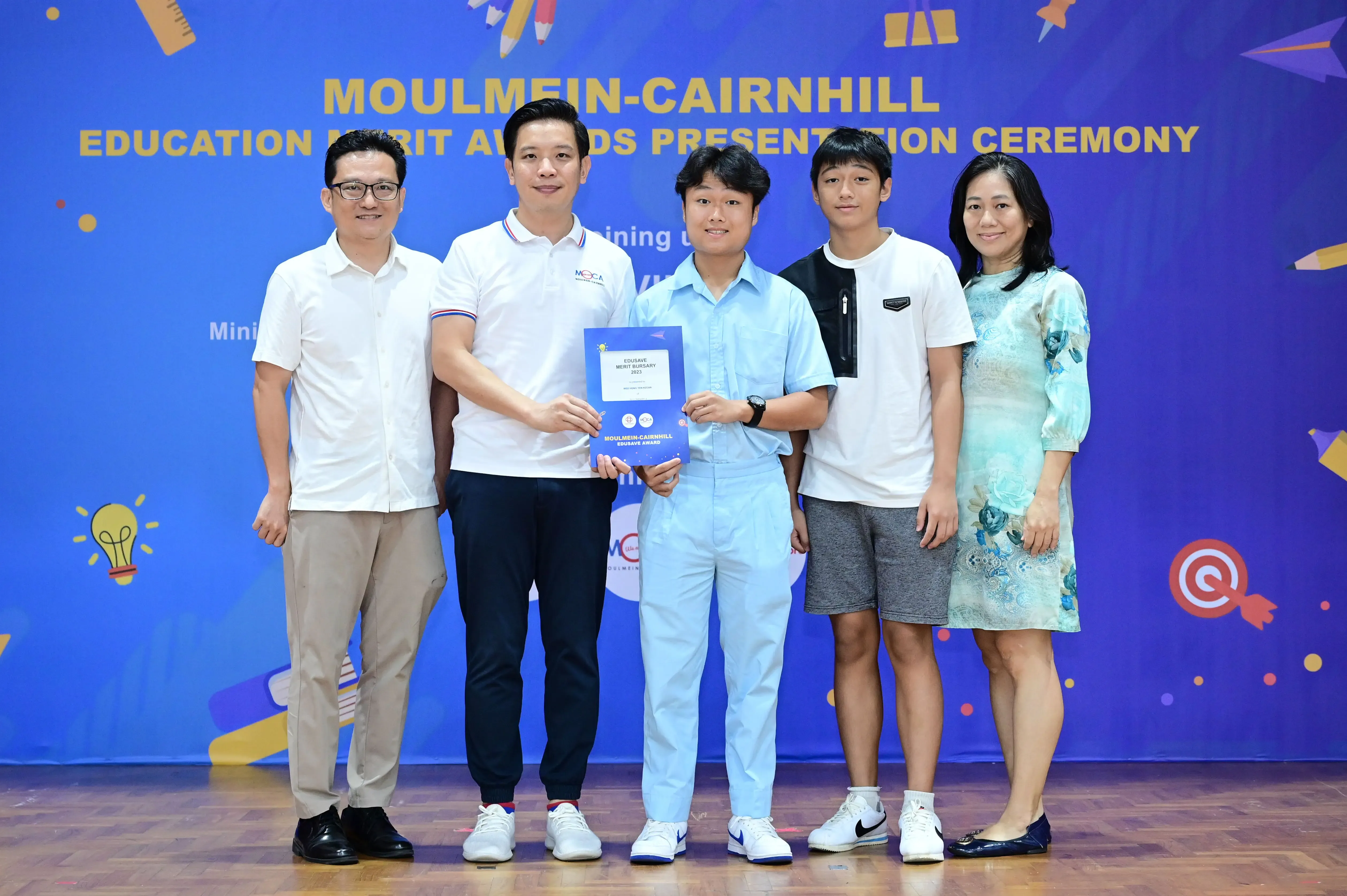 Five people pose at the Moulmein-Cairnhill Education Merit Awards ceremony, with a student holding a blue award certificate.