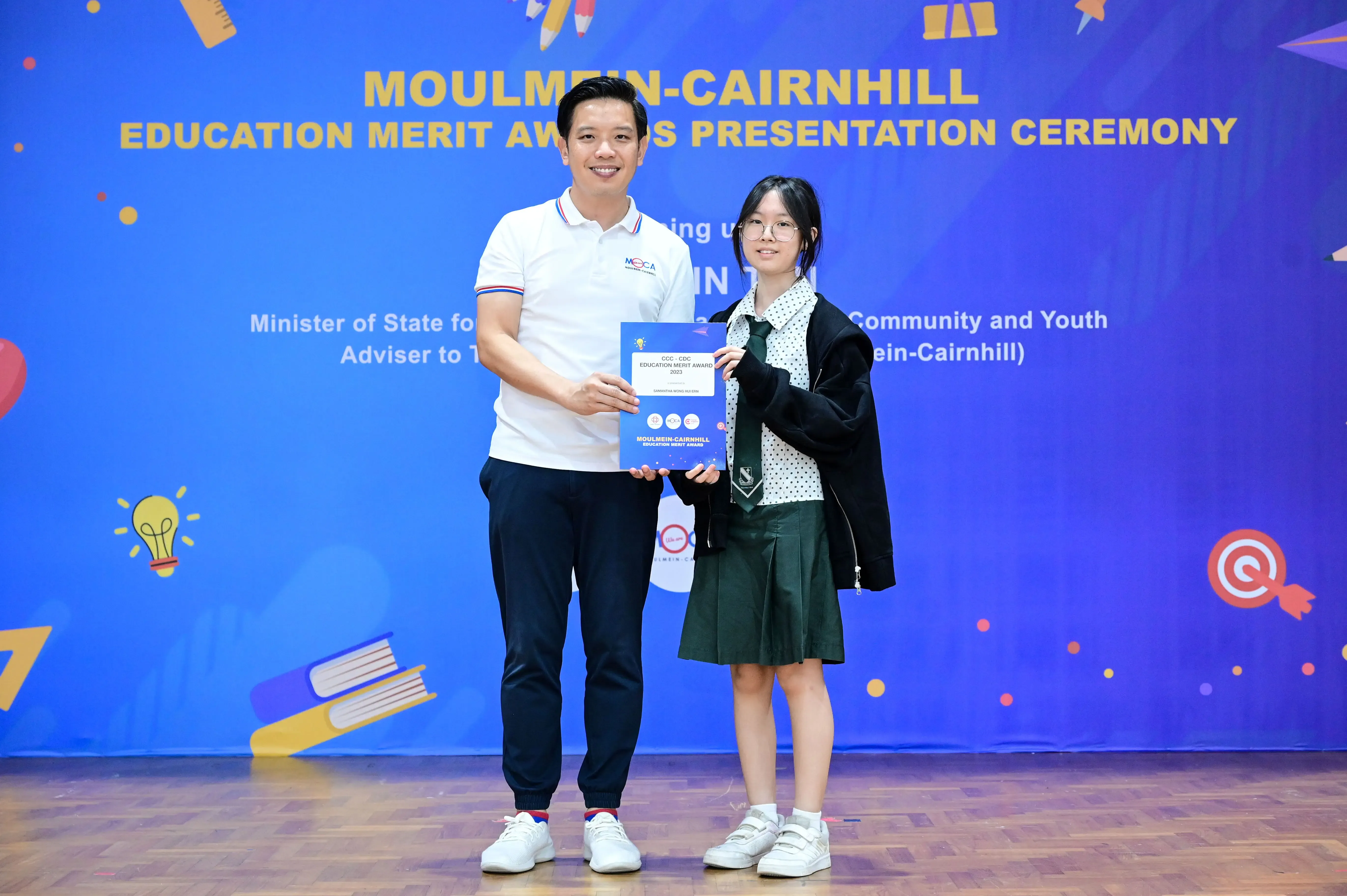 A man and a student in school uniform hold a certificate at the Moulmein-Cairnhill Education Merit Awards ceremony.
