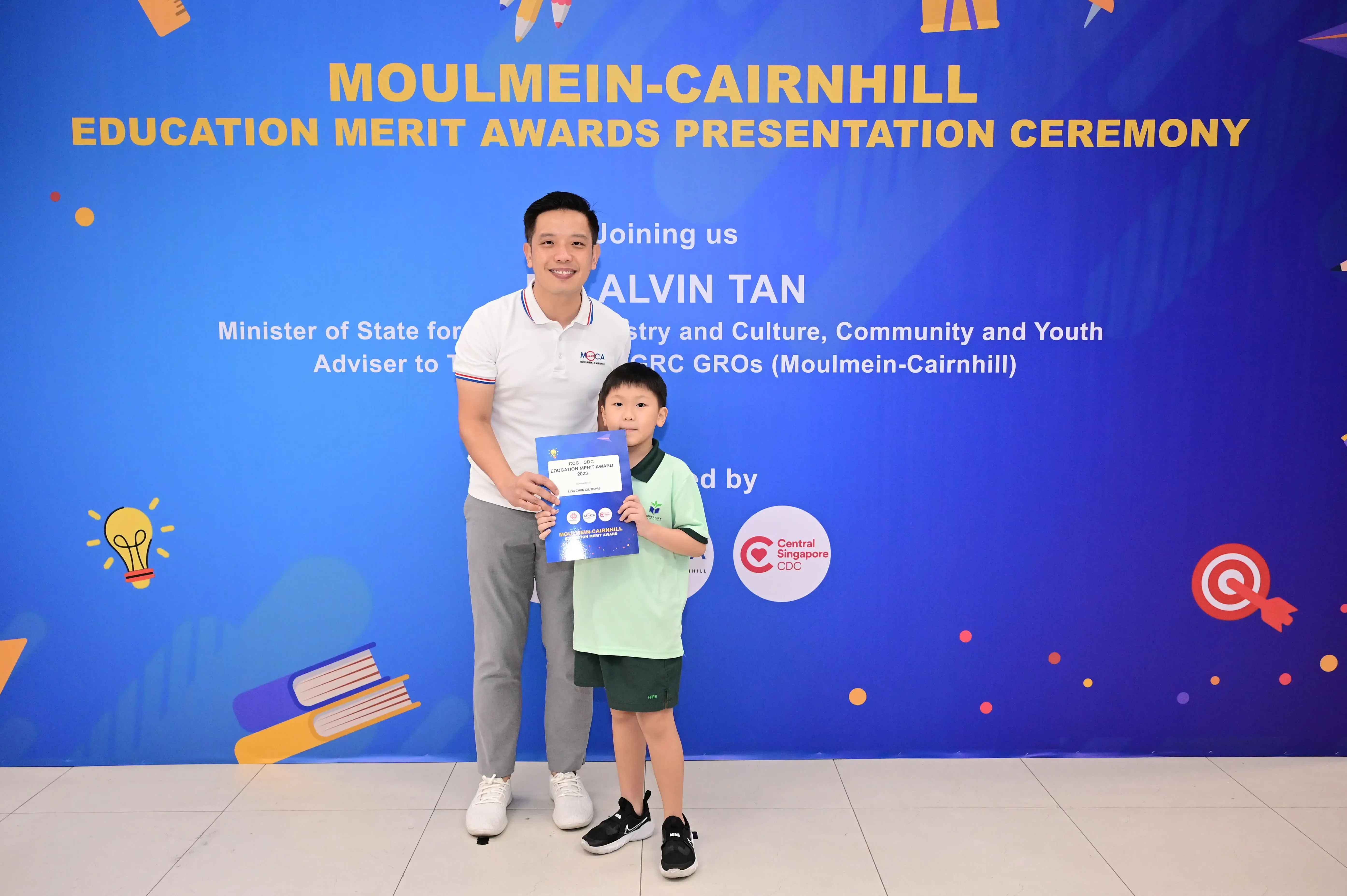A man and a young boy hold a certificate in front of a Moulmein-Cairnhill Education Merit Awards banner.