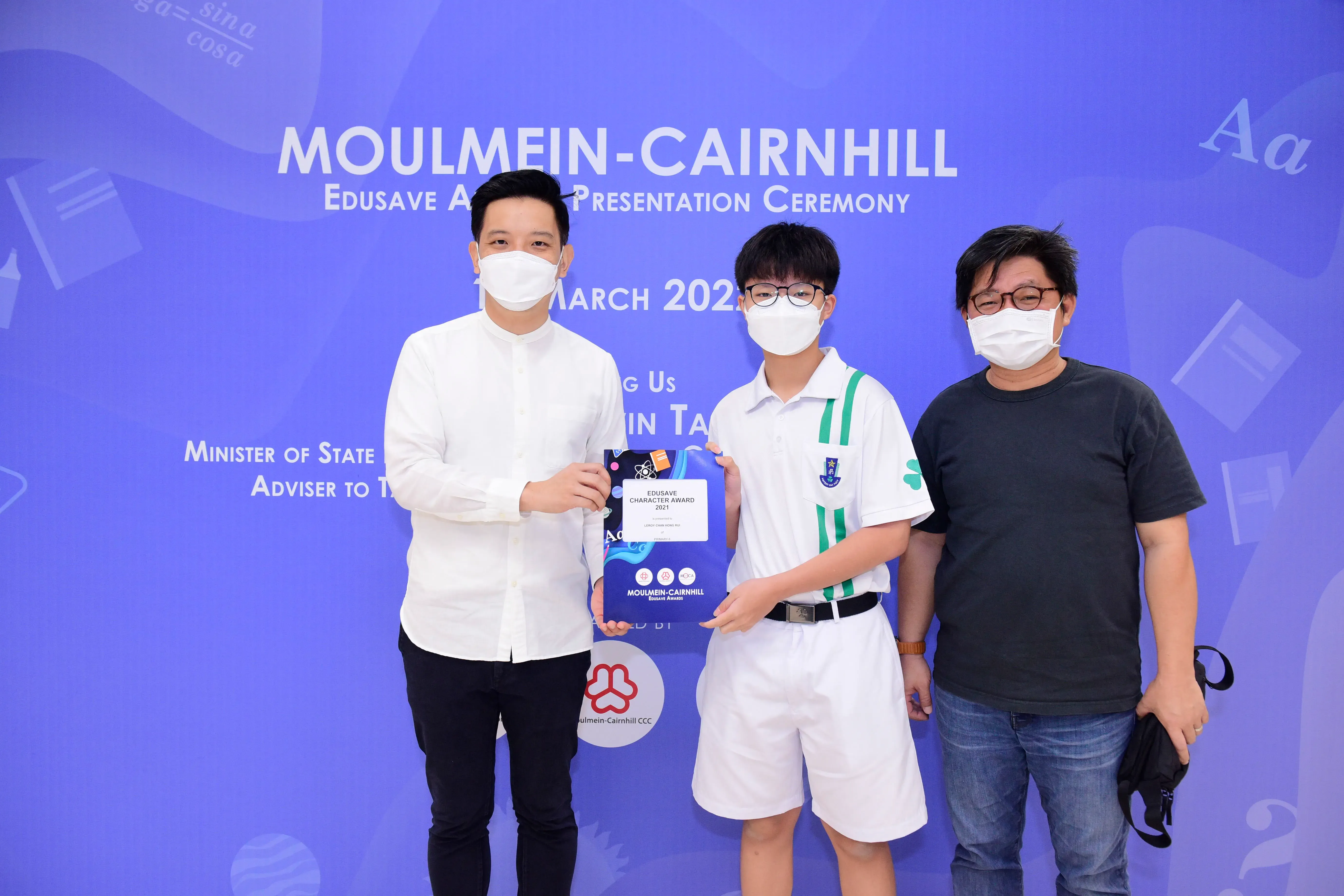 Three masked people pose at the Moulmein-Cairnhill Edusave Award Presentation Ceremony, with a student holding an award.
