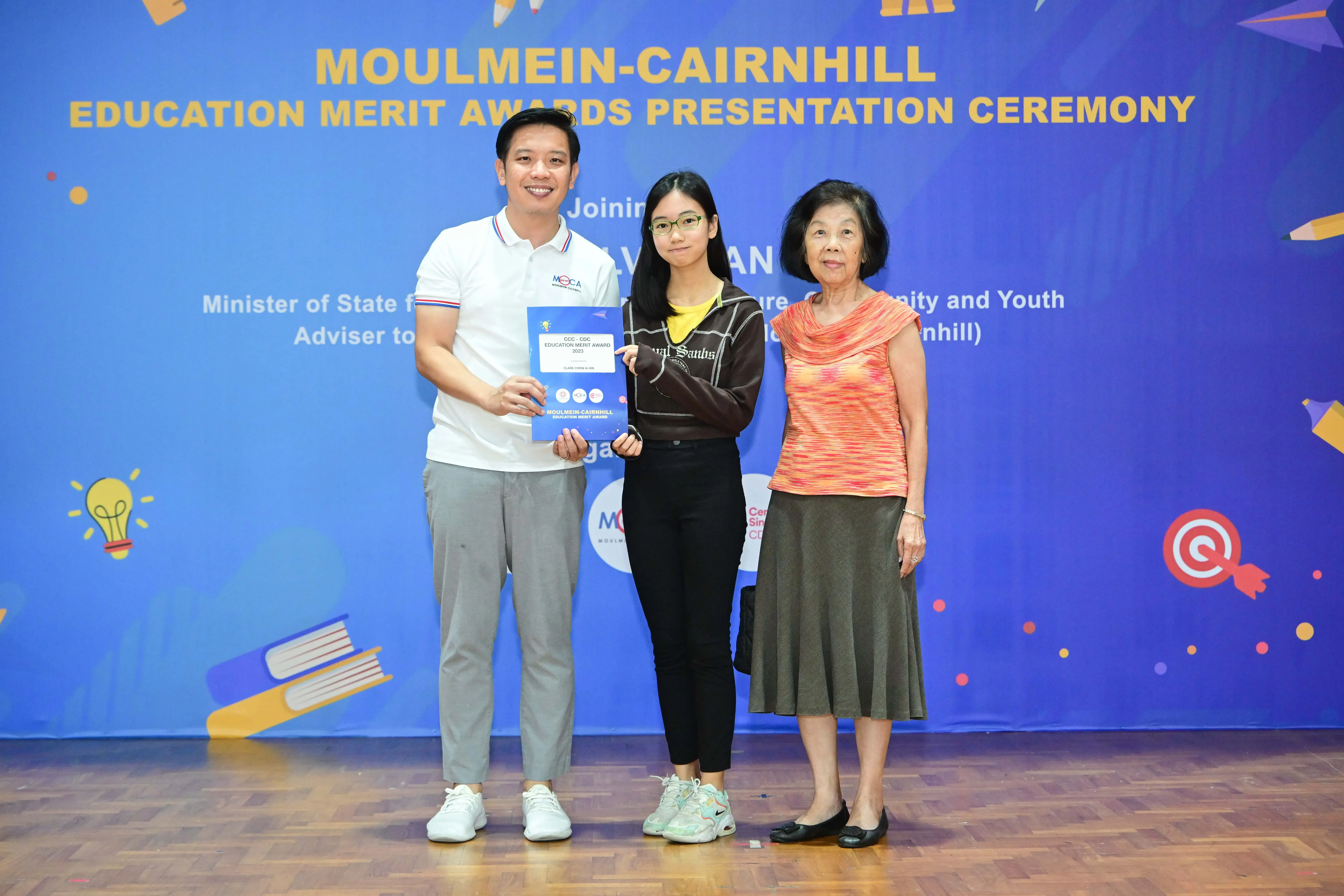 Three people pose together at the Moulmein-Cairnhill Education Merit Awards Presentation Ceremony, holding a certificate.