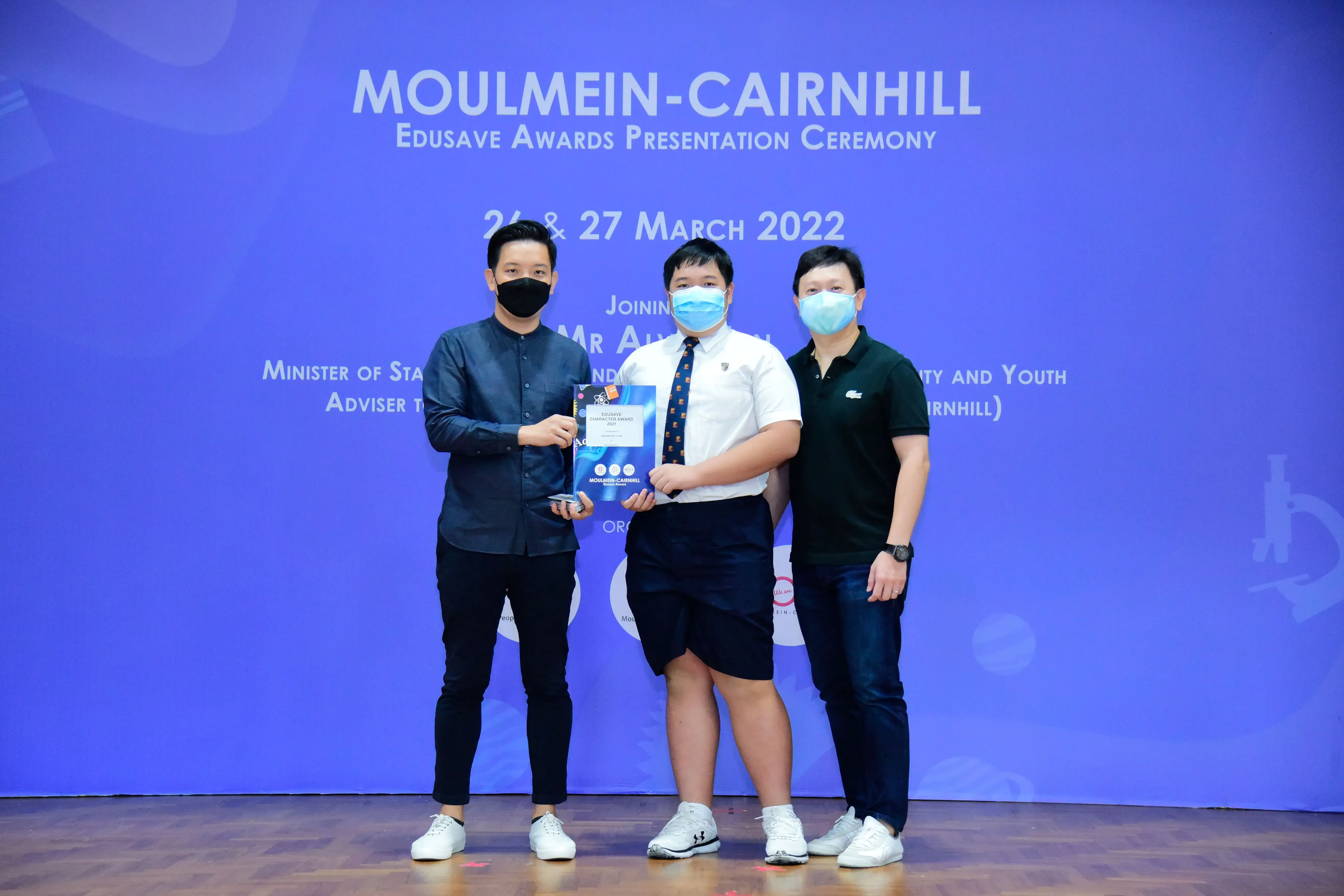 Three masked people pose with an award at the Moulmein-Cairnhill Edusave Awards Presentation Ceremony, March 2022.