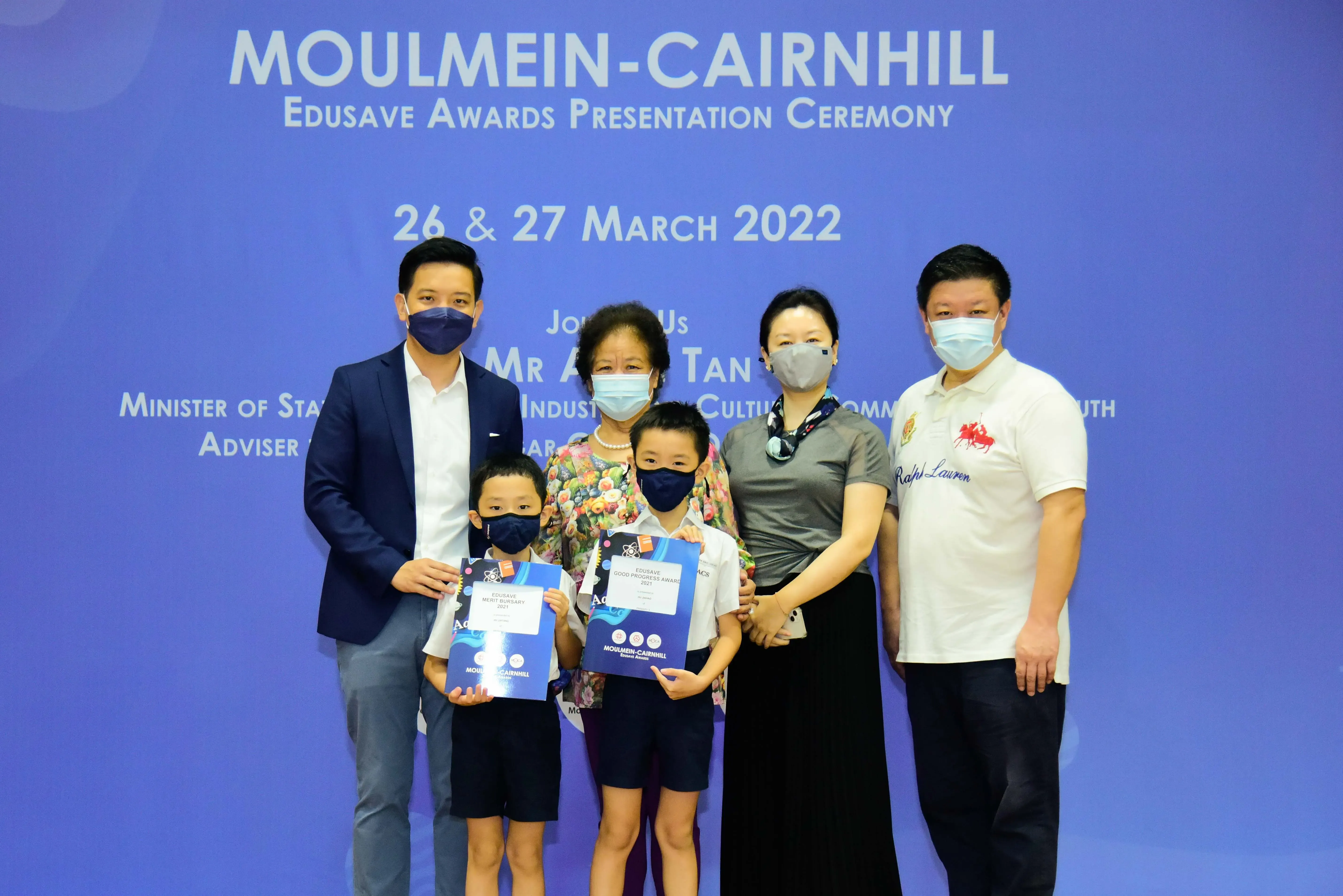 Five masked adults and two children holding award folders pose at the Moulmein-Cairnhill Edusave Awards Ceremony, March 2022.