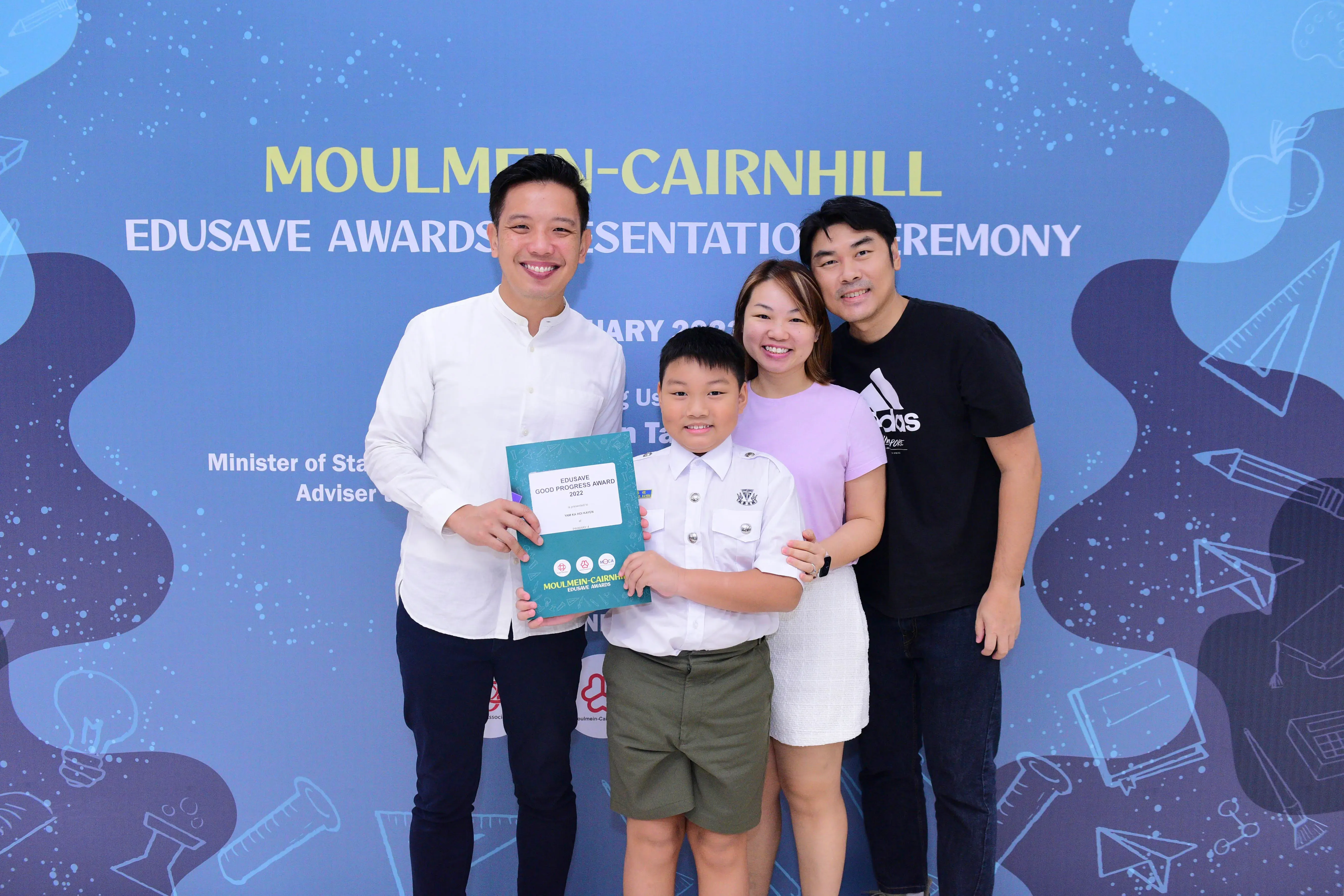 Four people smile at the Moulmein-Cairnhill Edusave Awards Ceremony, with a boy holding a 2022 Good Progress Award certificate.