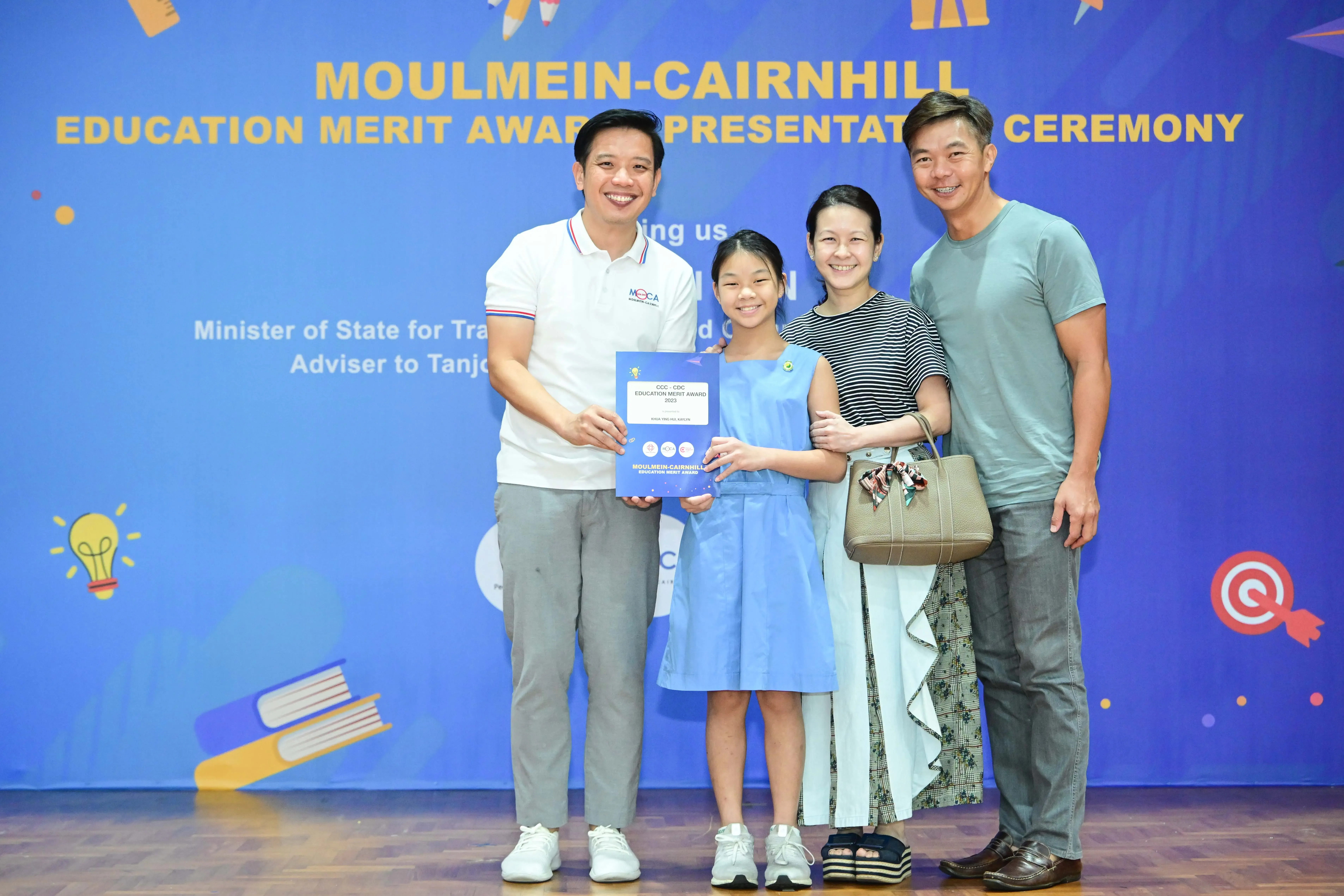 Four people pose smiling at the Moulmein-Cairnhill Education Merit Award Presentation Ceremony, with a girl holding a certificate.