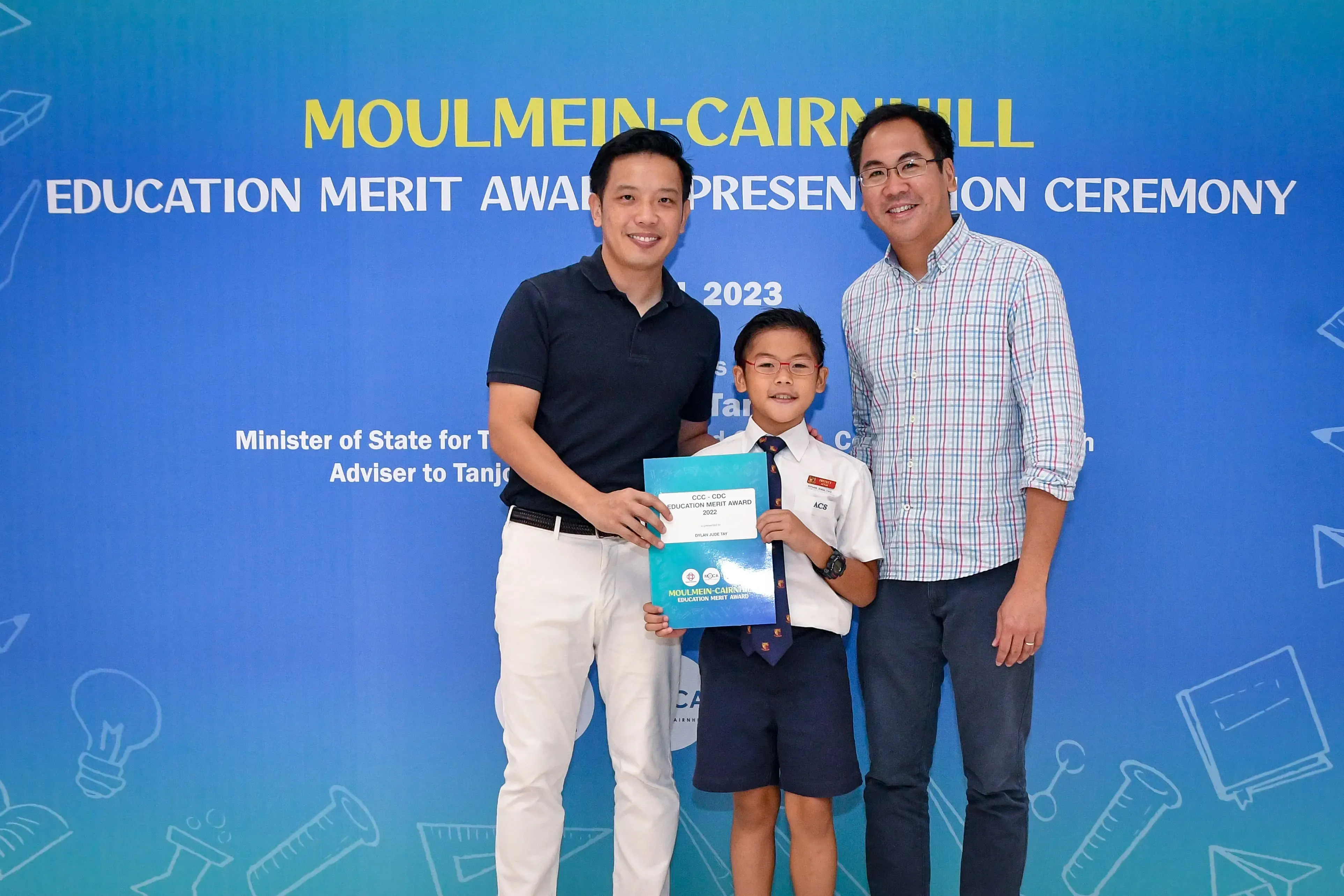 Two men and a boy in school uniform hold an award certificate at the 2023 Moulmein-Cairnhill Education Merit Award ceremony.