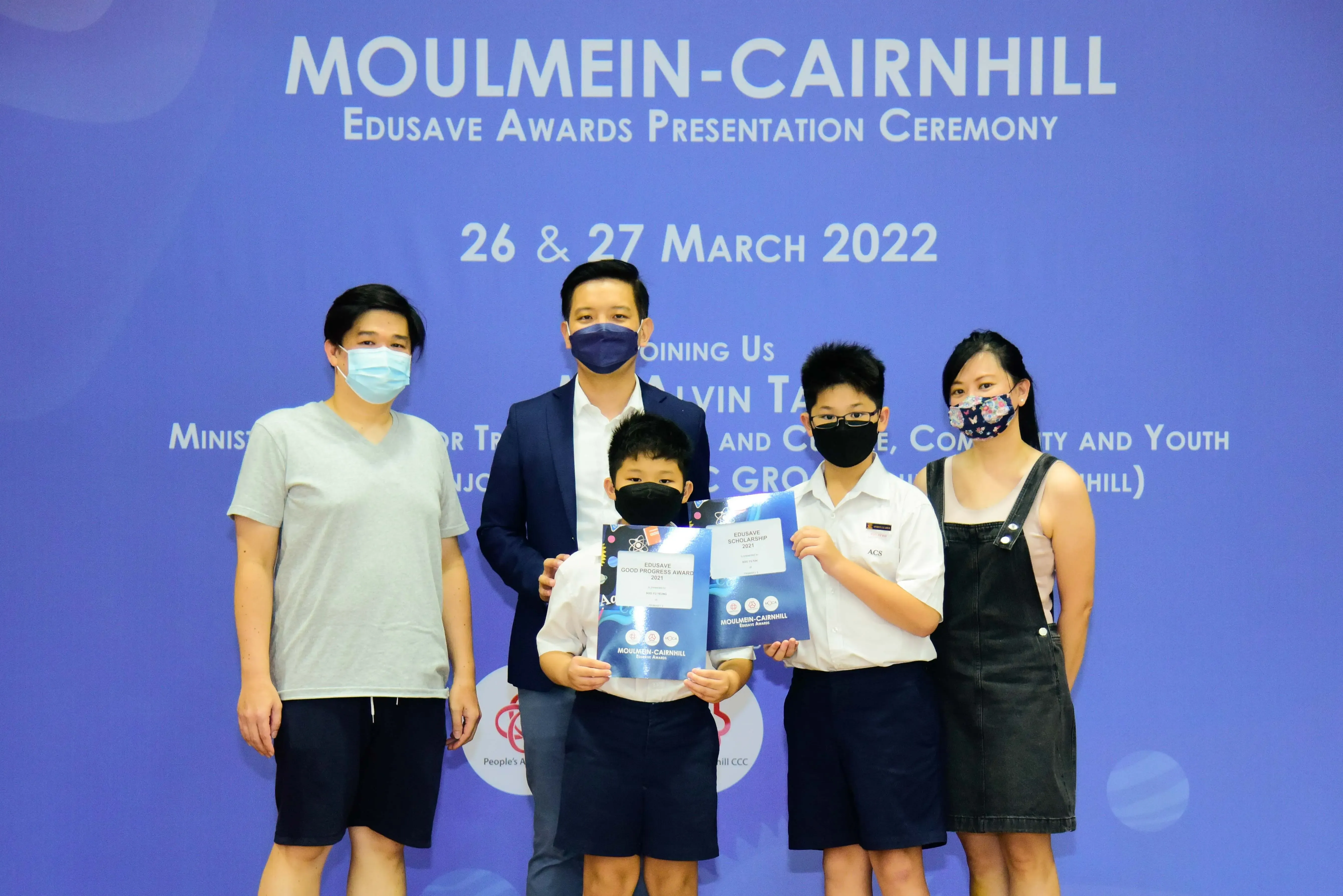 Five masked people pose at the Moulmein-Cairnhill Edusave Awards Ceremony; two children hold award certificates.