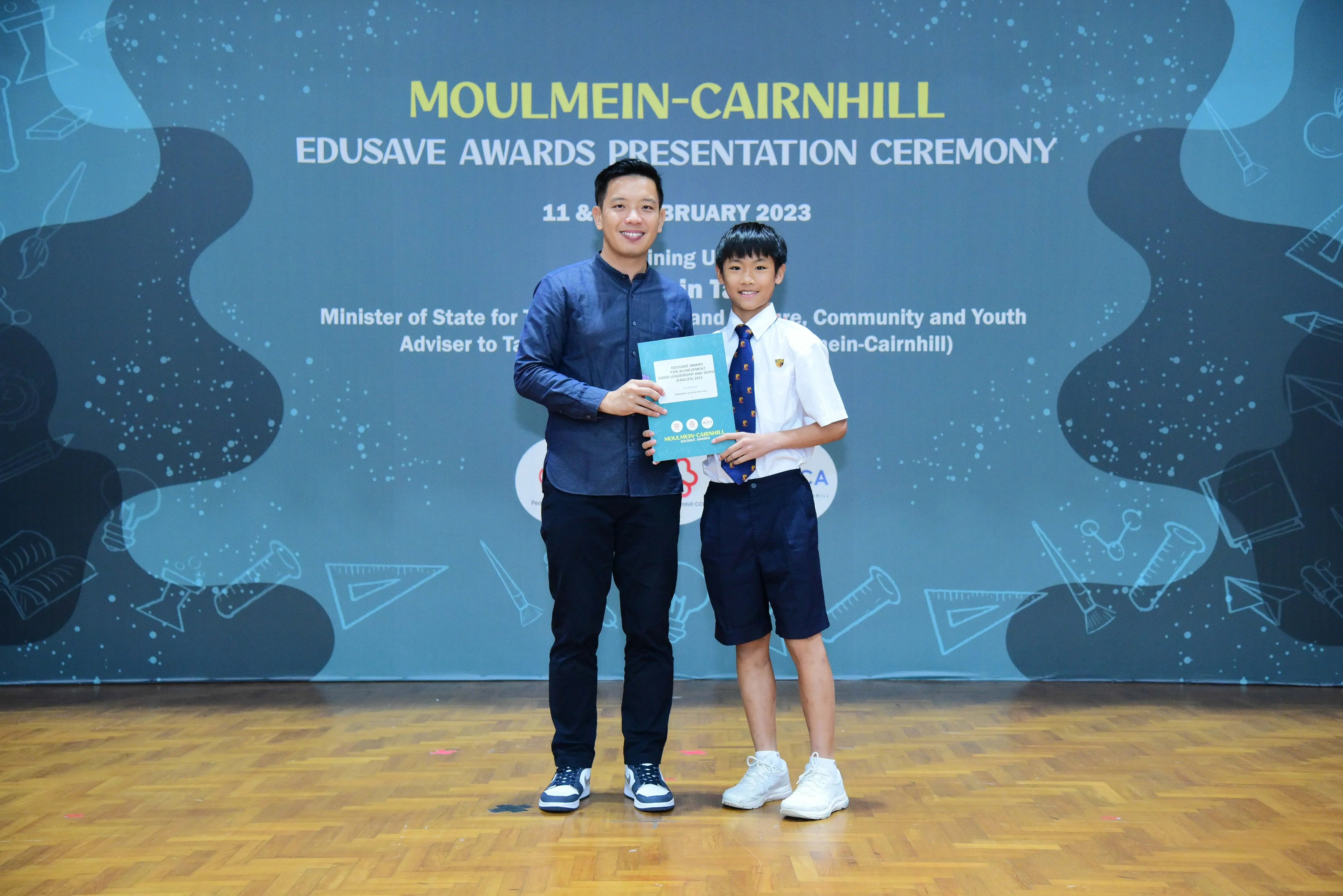 An adult and a young student in uniform hold a certificate at the Moulmein-Cairnhill Edusave Awards Ceremony 2023.