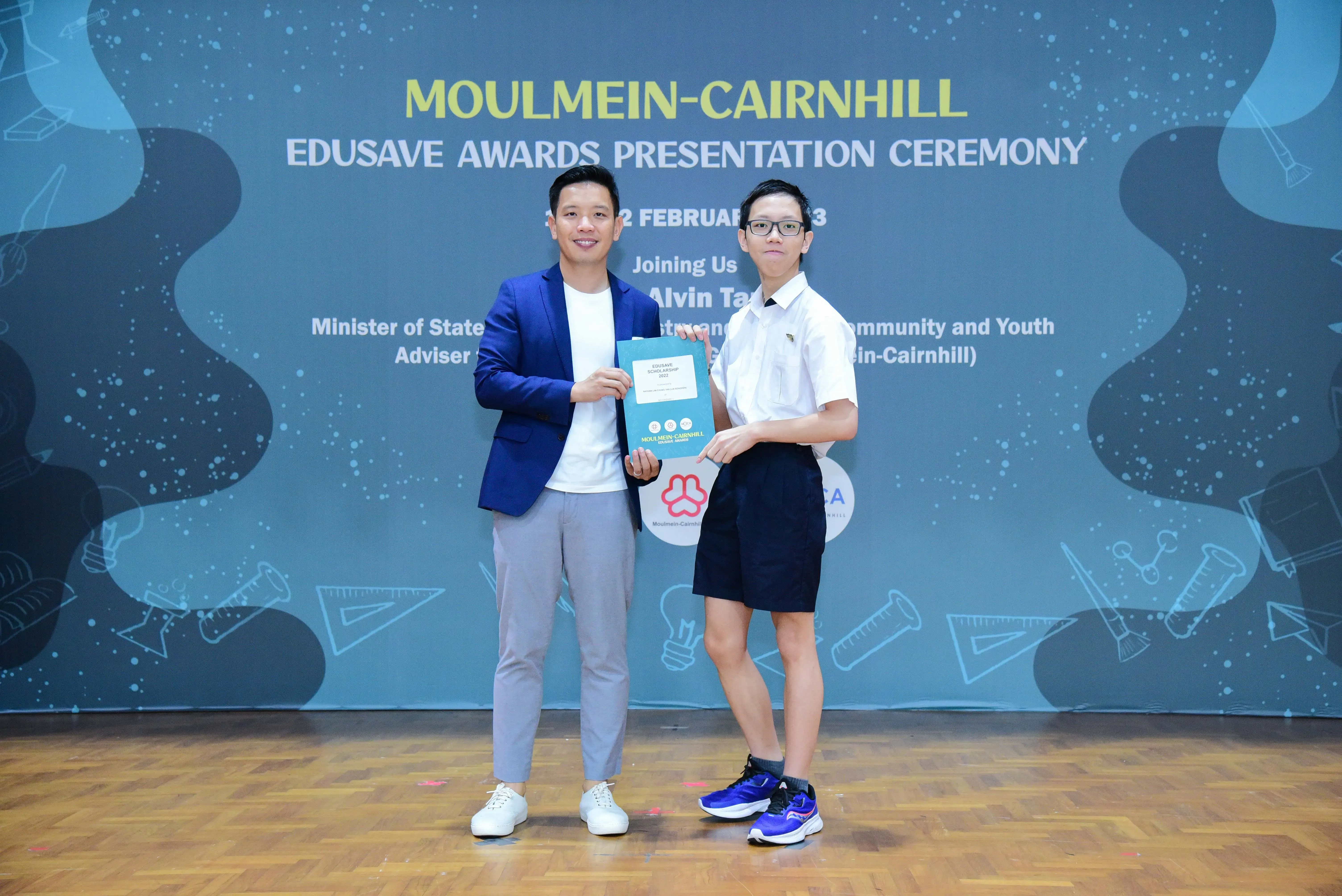 Two people hold a certificate together on stage at the Moulmein-Cairnhill Edusave Awards Presentation Ceremony.