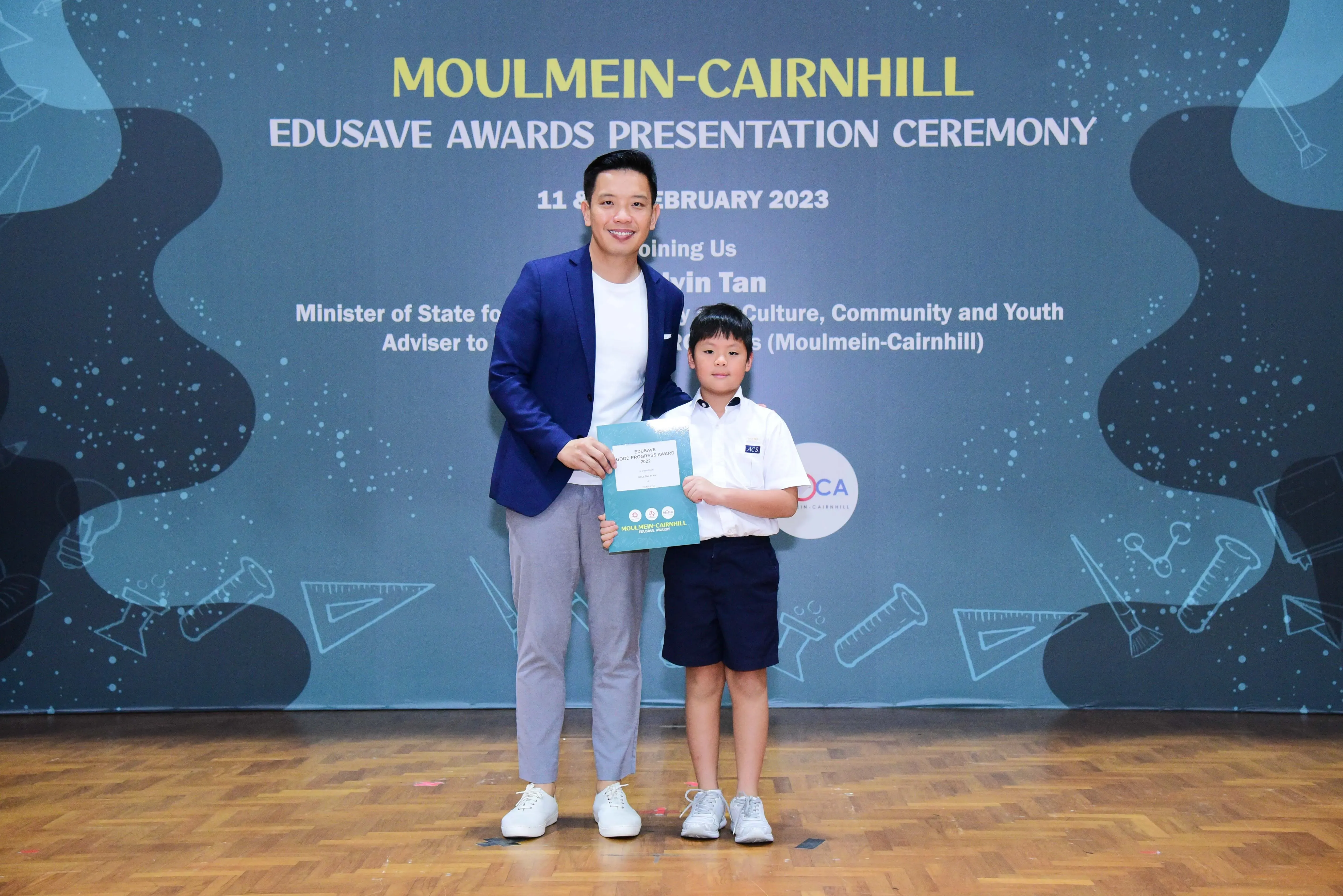 A man in a blue blazer and a young boy in school uniform hold a certificate at the Moulmein-Cairnhill Edusave Awards 2023.