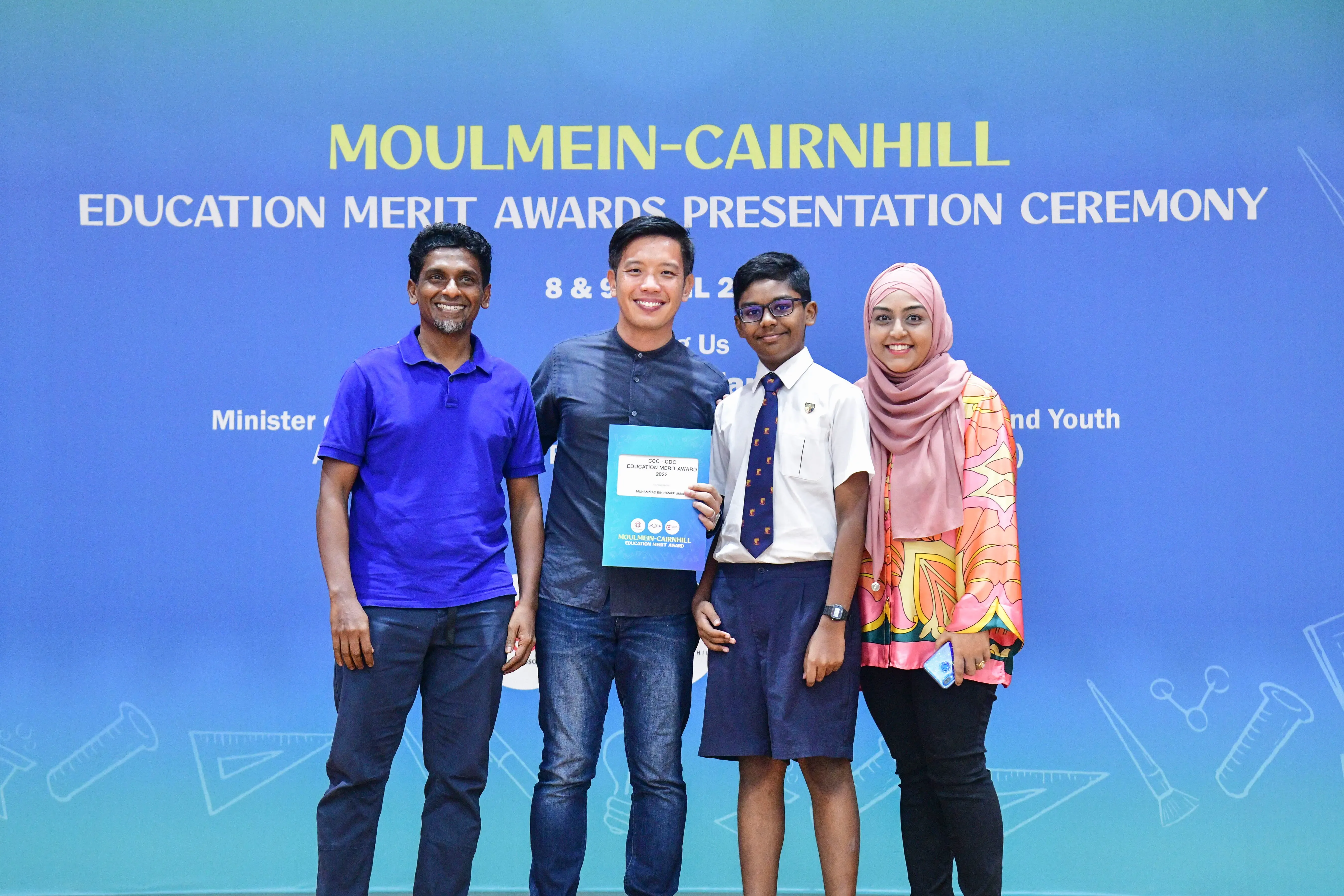 Four people pose smiling at the Moulmein-Cairnhill Education Merit Awards Presentation Ceremony, one holding a blue award folder.