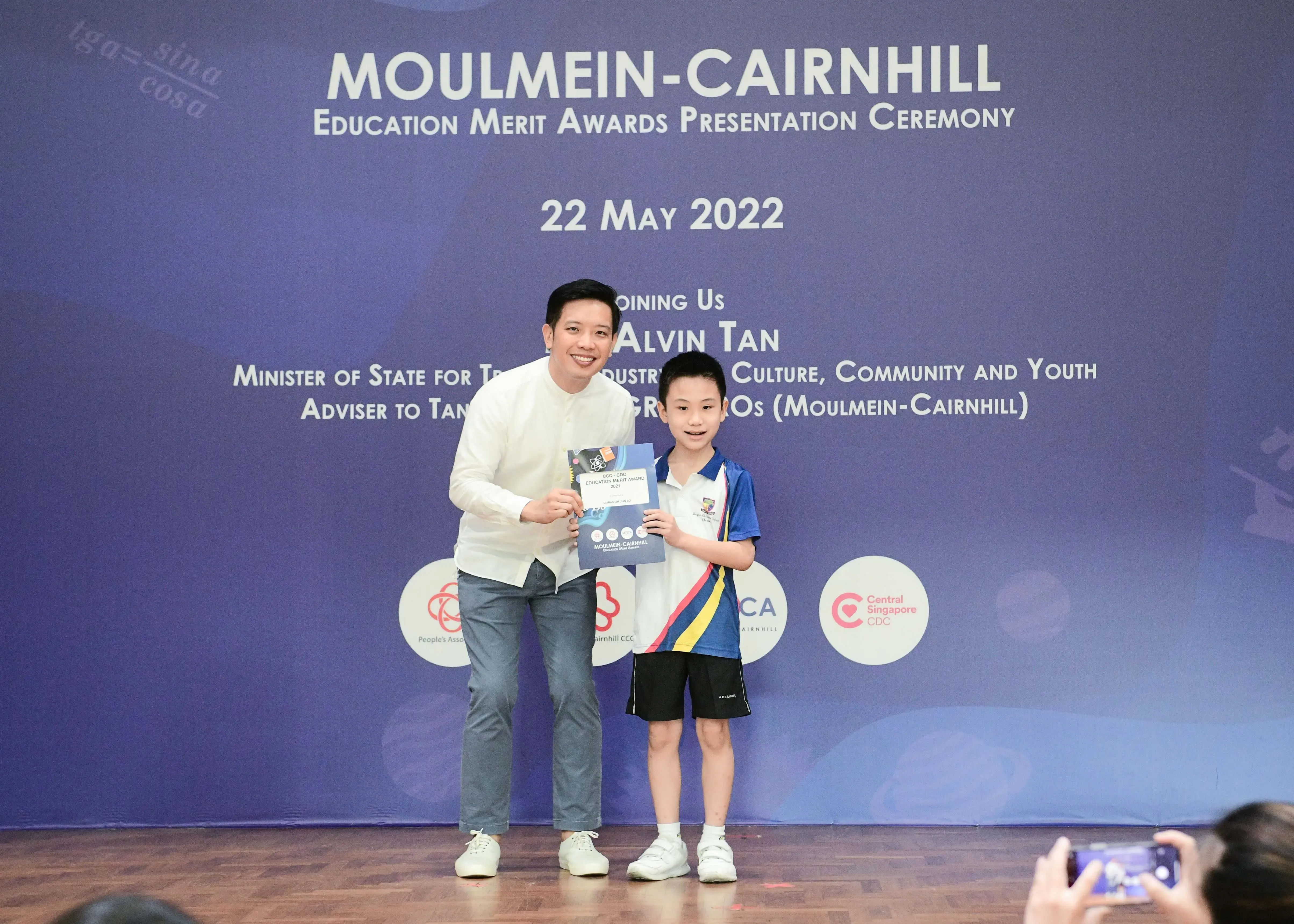 A man and a young boy hold a certificate on stage at the Moulmein-Cairnhill Education Merit Awards, 22 May 2022.