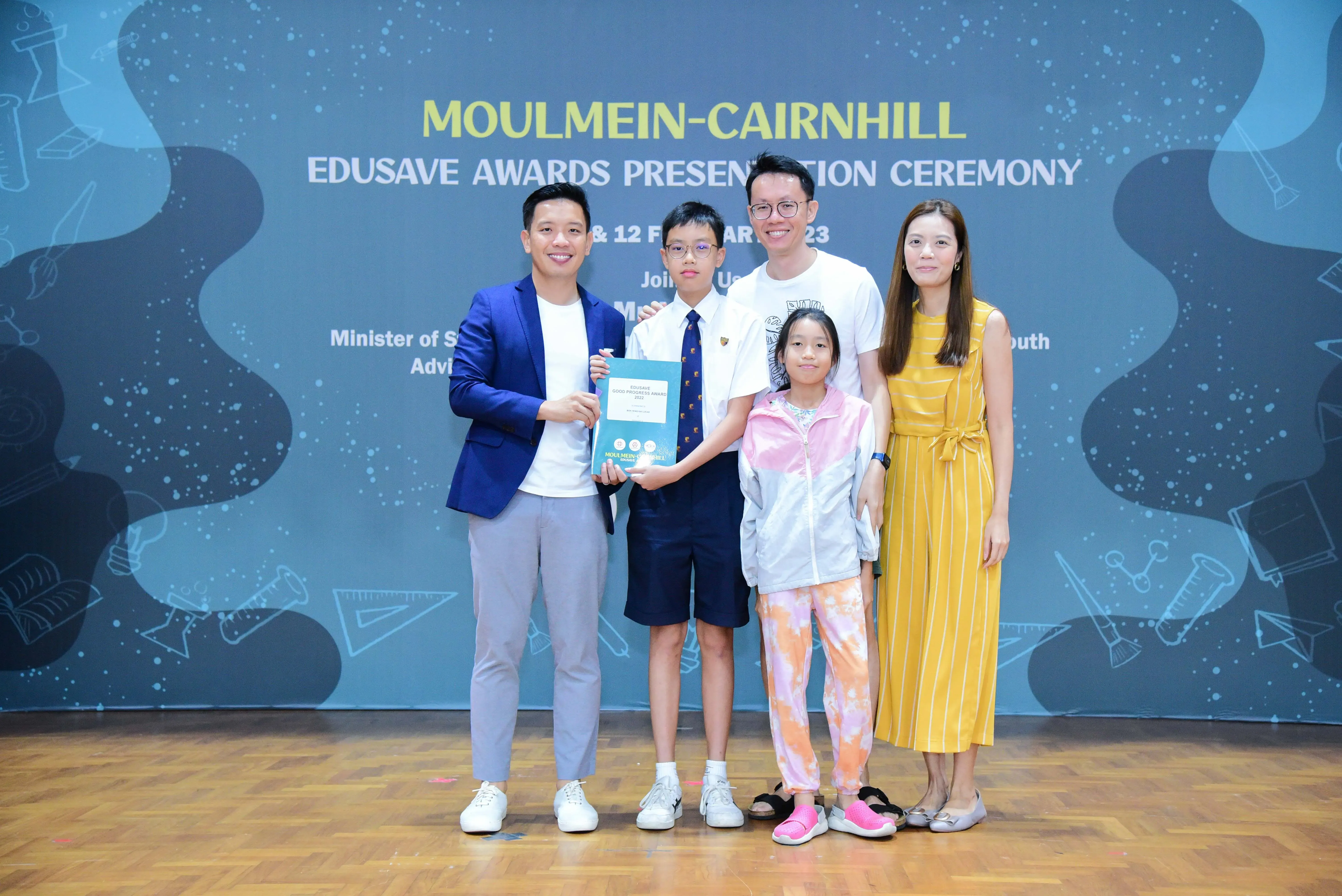 Five people pose with an award booklet at the Moulmein-Cairnhill Edusave Awards Presentation Ceremony backdrop.