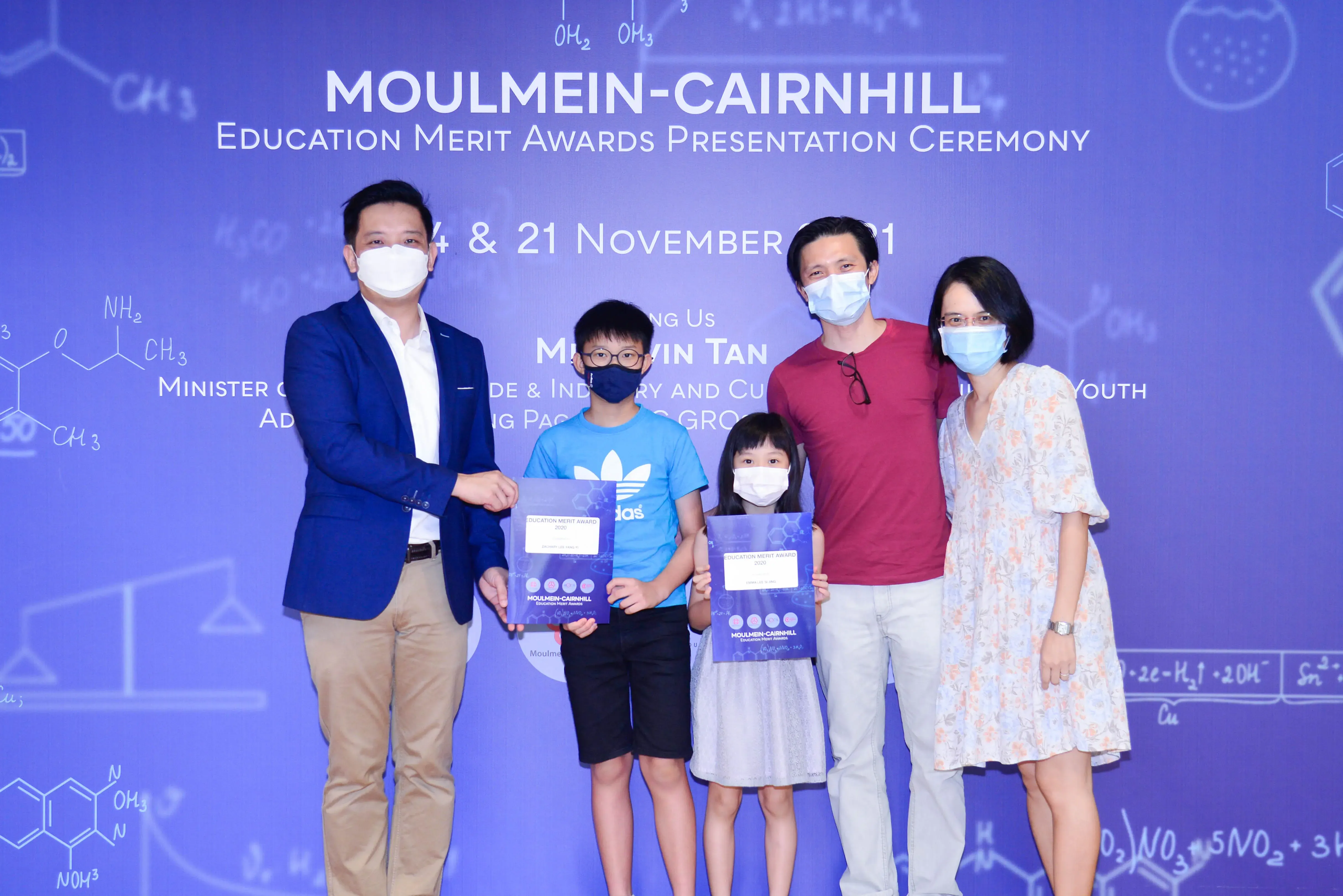 Five masked people pose at Moulmein-Cairnhill Education Merit Awards Presentation Ceremony, two children holding award certificates.