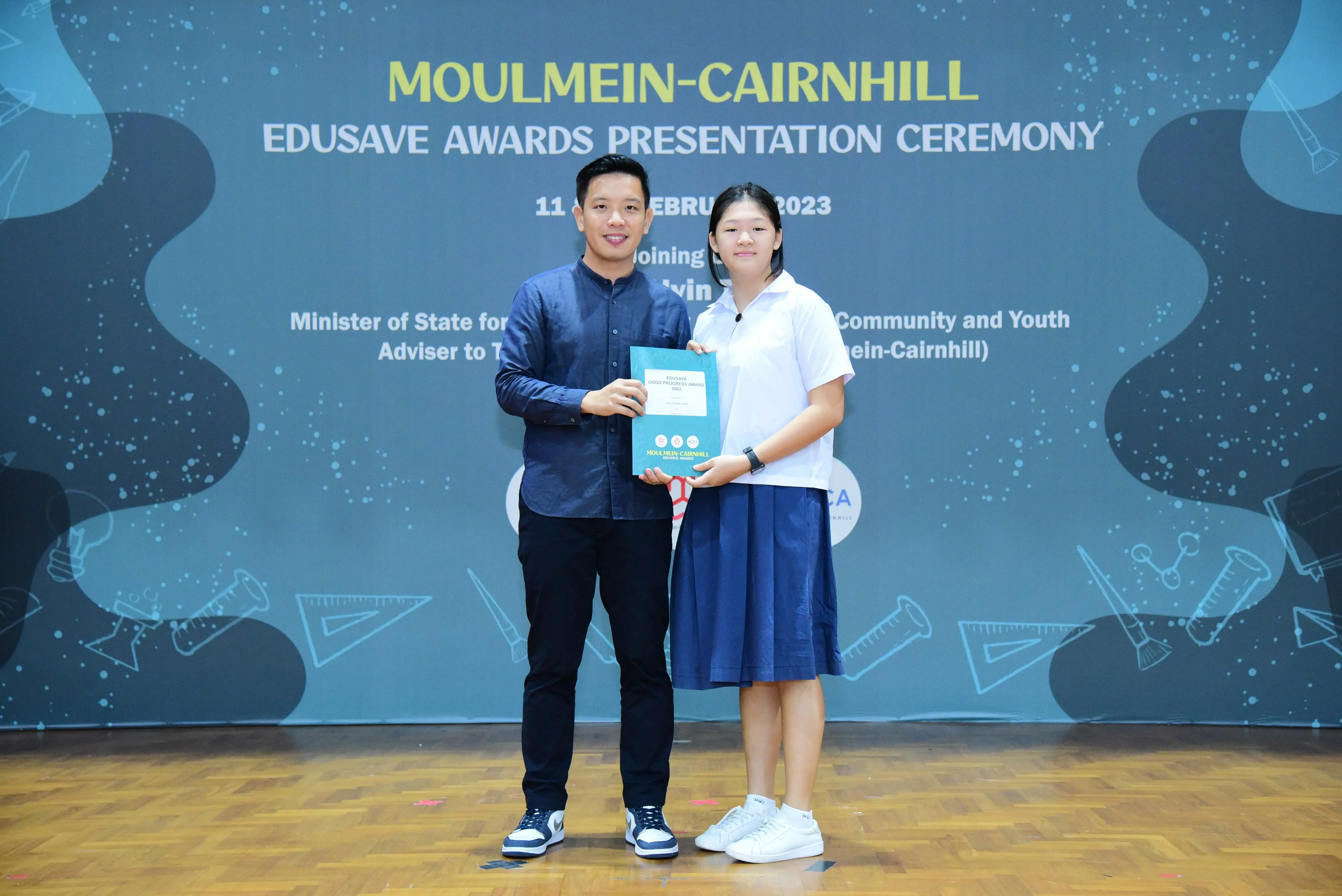Two people hold a certificate together on stage at the Moulmein-Cairnhill Edusave Awards Presentation Ceremony 2023.
