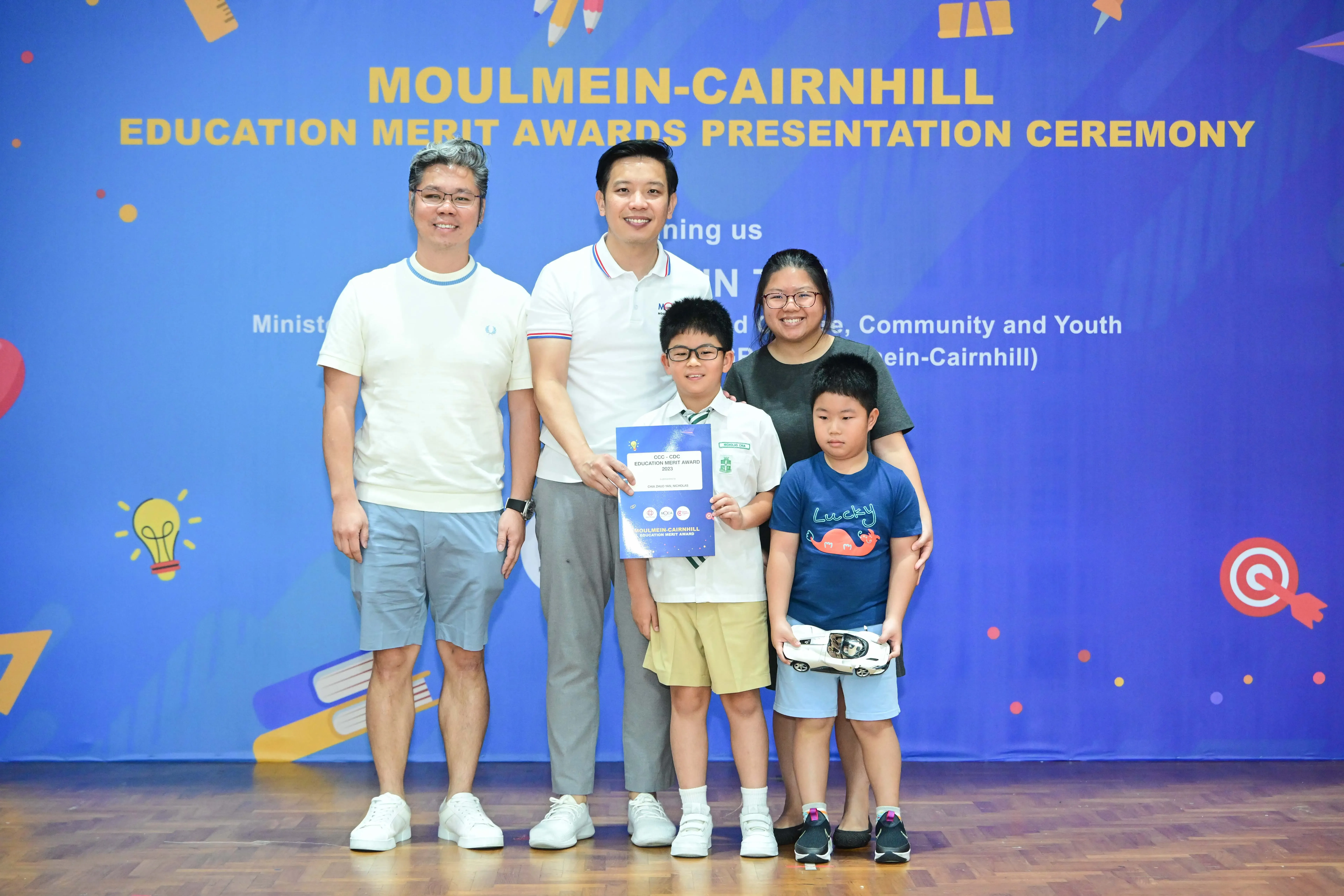 Five people pose before a Moulmein-Cairnhill Education Merit Awards banner; a boy holds a certificate.