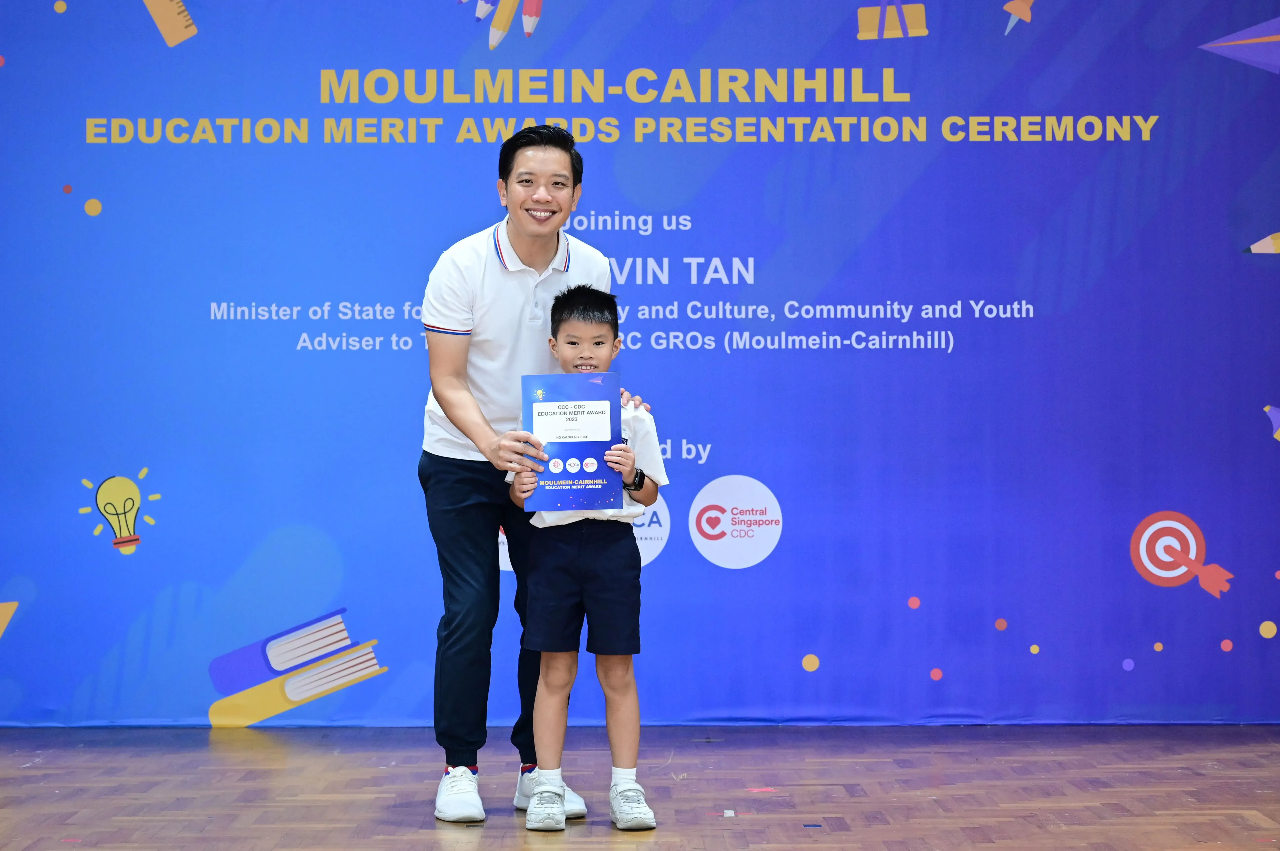 A man and young boy hold a blue certificate at the Moulmein-Cairnhill Education Merit Awards Presentation Ceremony.