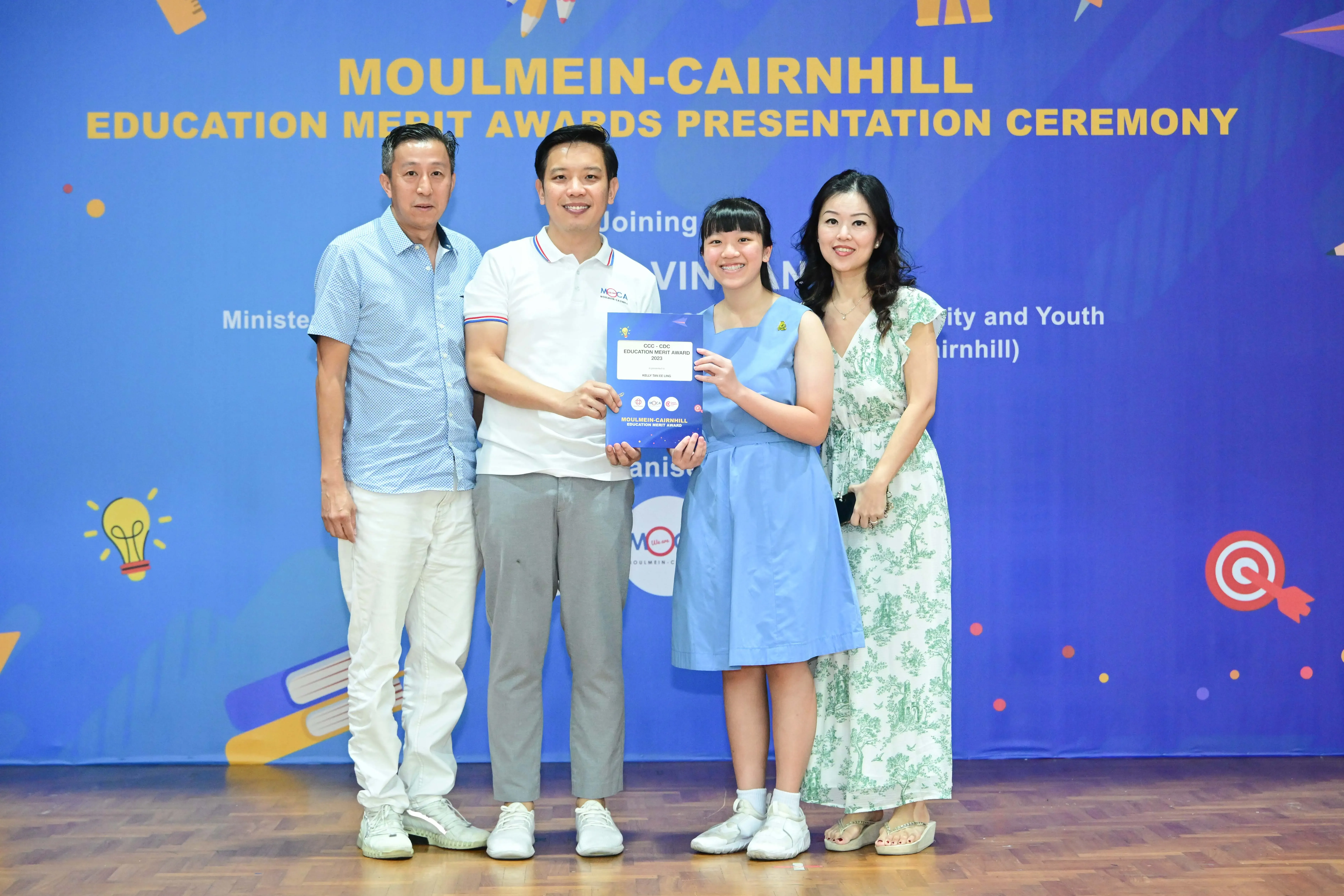 Four people pose together at the Moulmein-Cairnhill Education Merit Awards, with a young girl holding a certificate.
