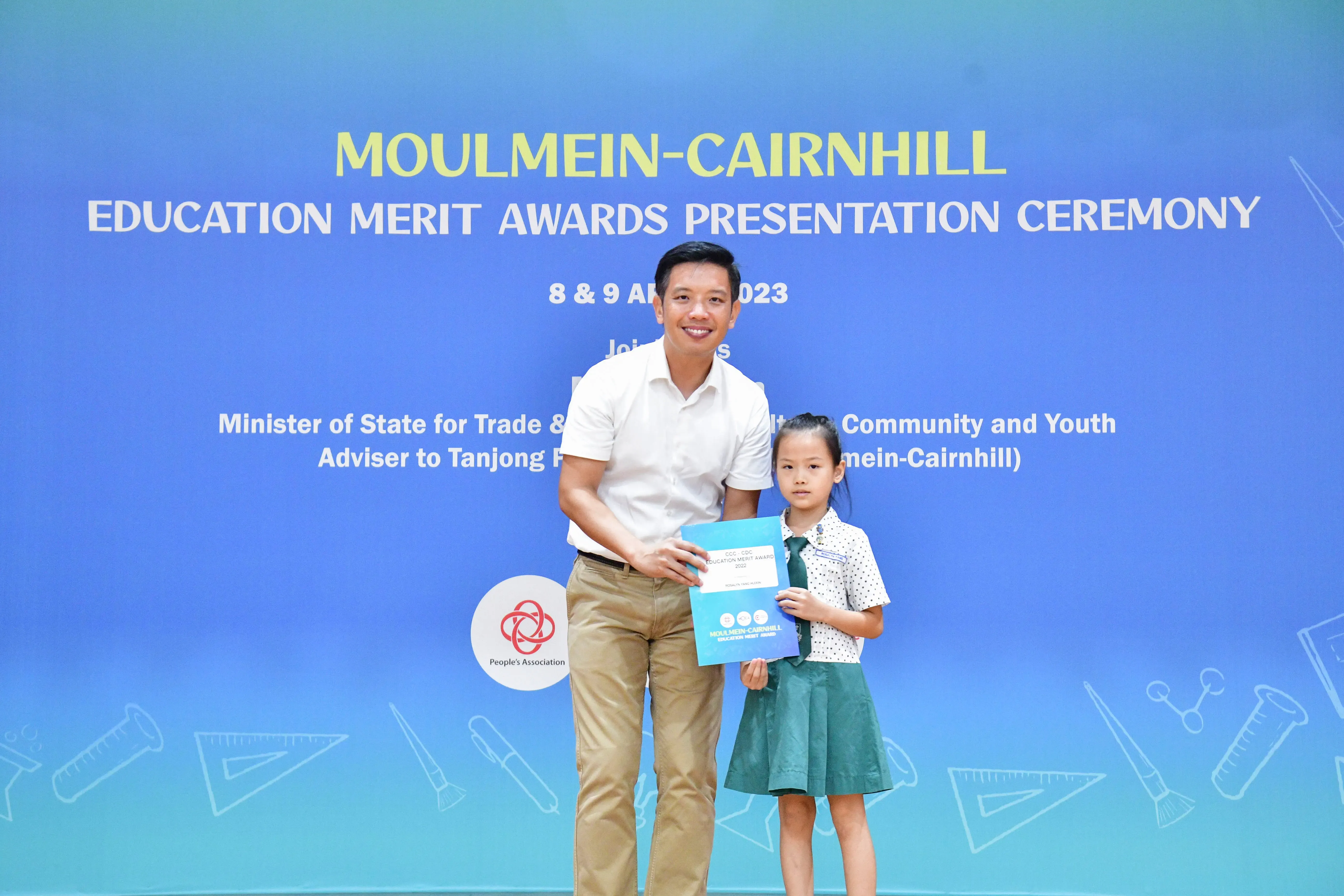 A man and a young girl hold an award certificate in front of a Moulmein-Cairnhill Education Merit Awards banner.