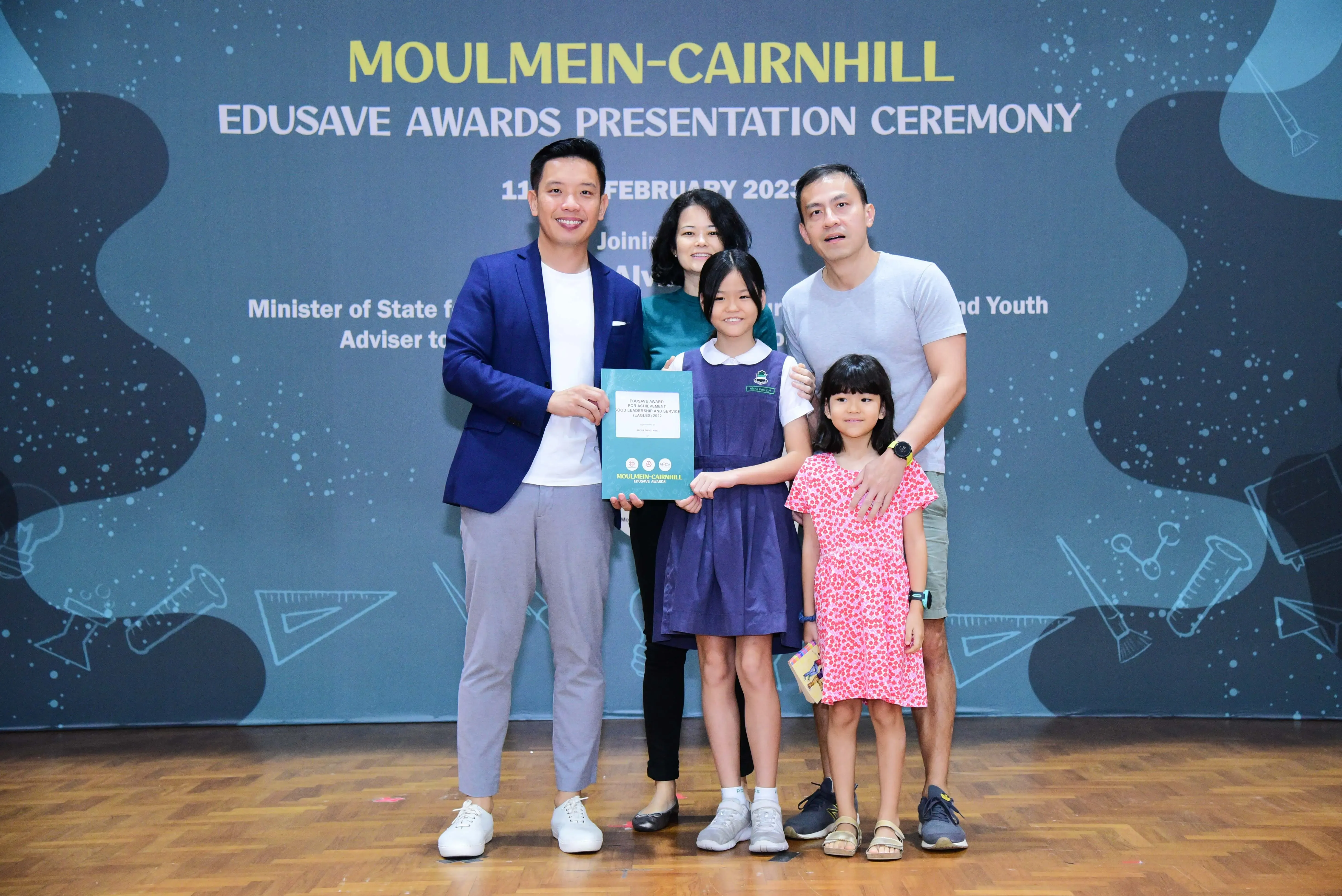Five people pose with an award certificate at the Moulmein-Cairnhill Edusave Awards Presentation Ceremony backdrop.