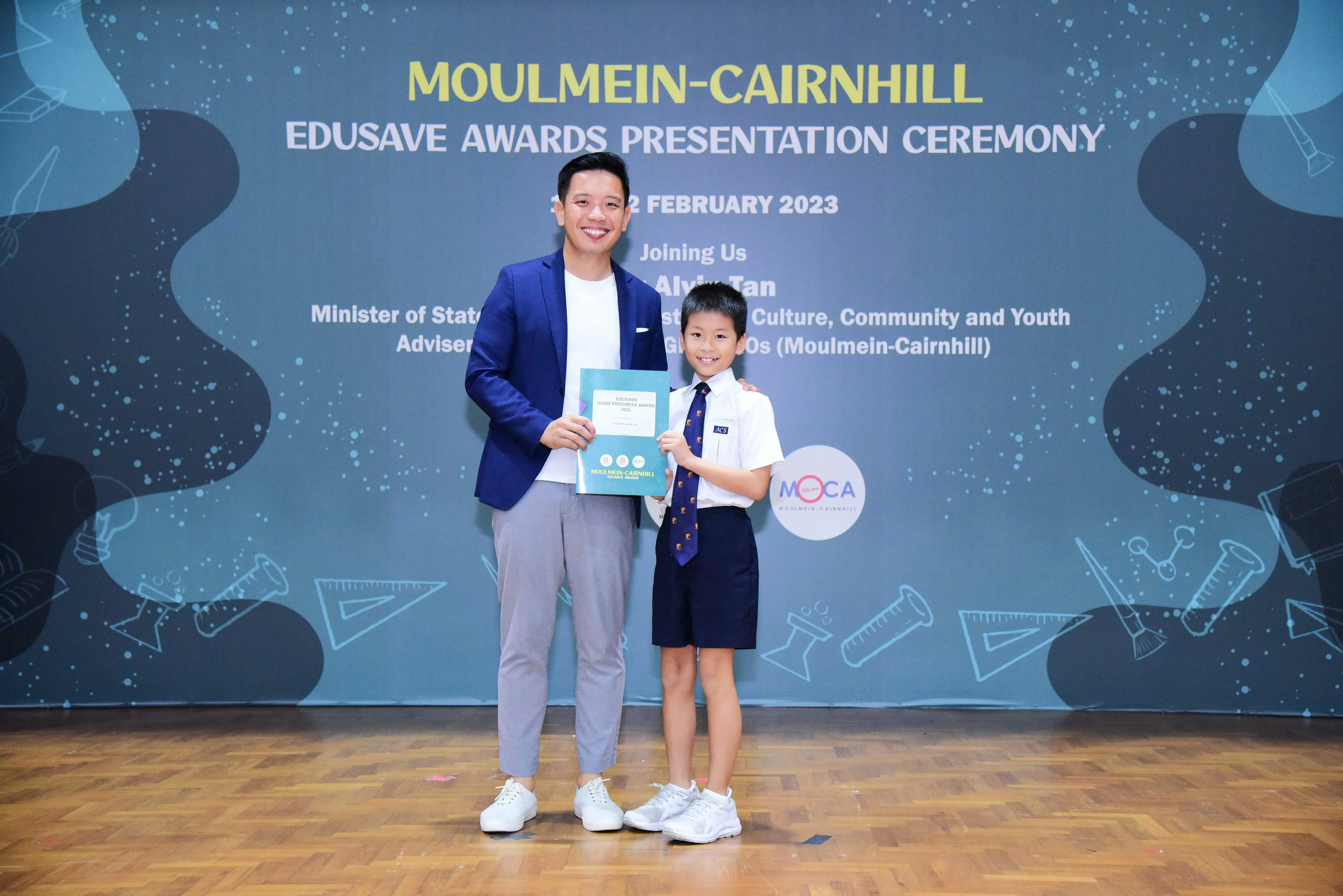 A man and a young boy in school uniform hold a certificate at the Moulmein-Cairnhill Edusave Awards Ceremony, February 2023.