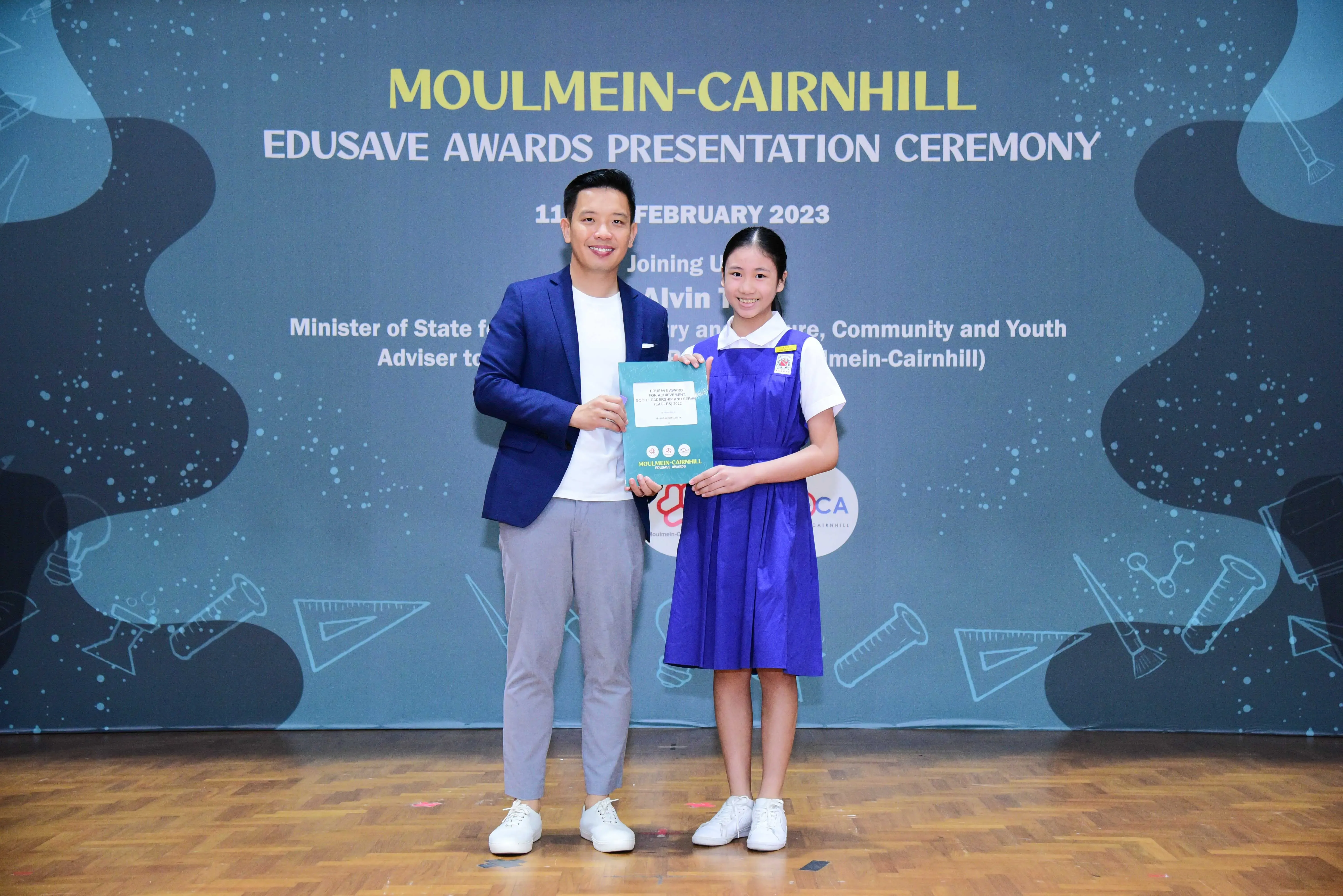 A man in a blue blazer and a young girl in a blue school uniform hold a certificate at the Moulmein-Cairnhill Edusave Awards Ceremony.