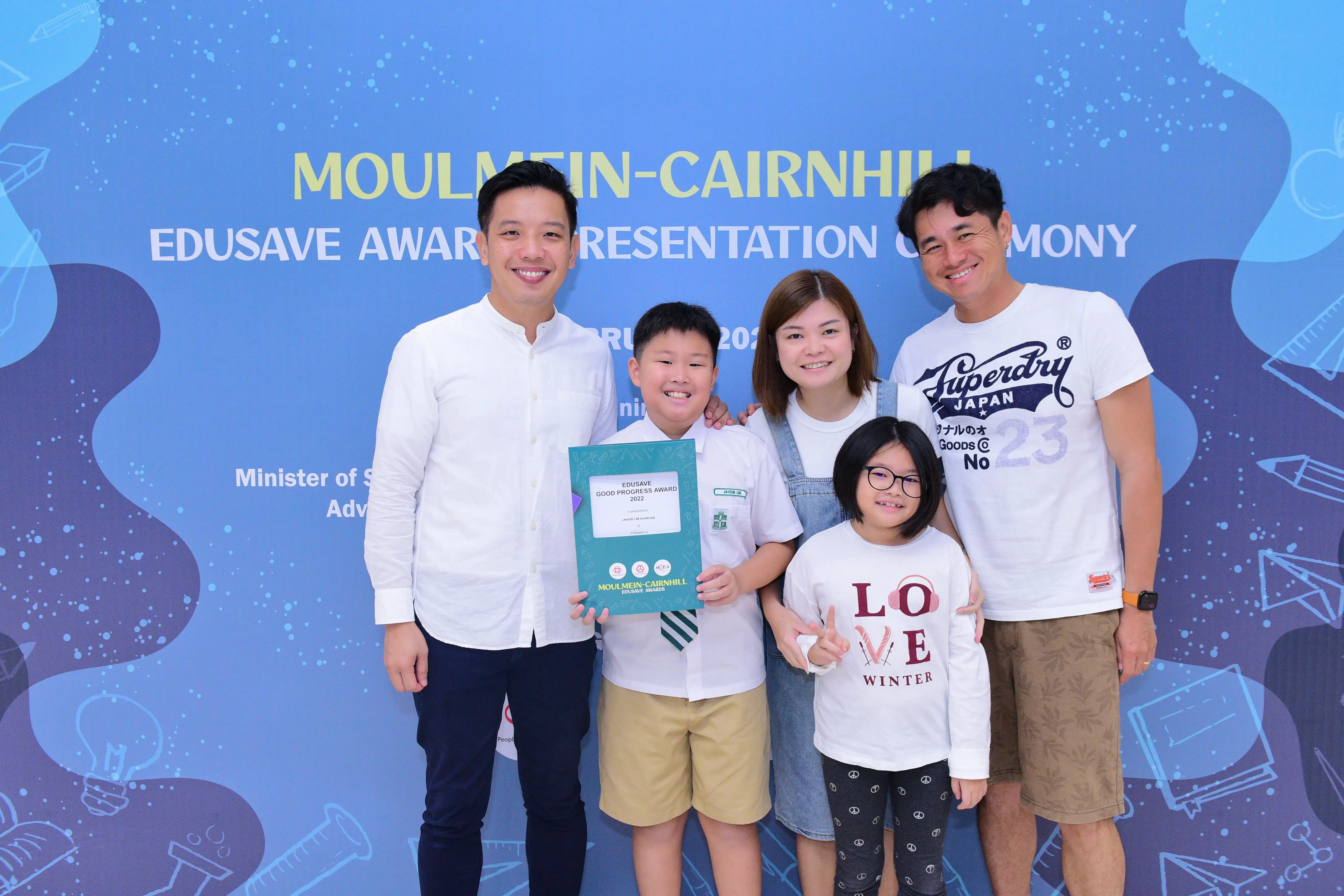 Five smiling people pose before a Moulmein-Cairnhill Edusave Award Presentation Ceremony banner, boy holding a certificate.