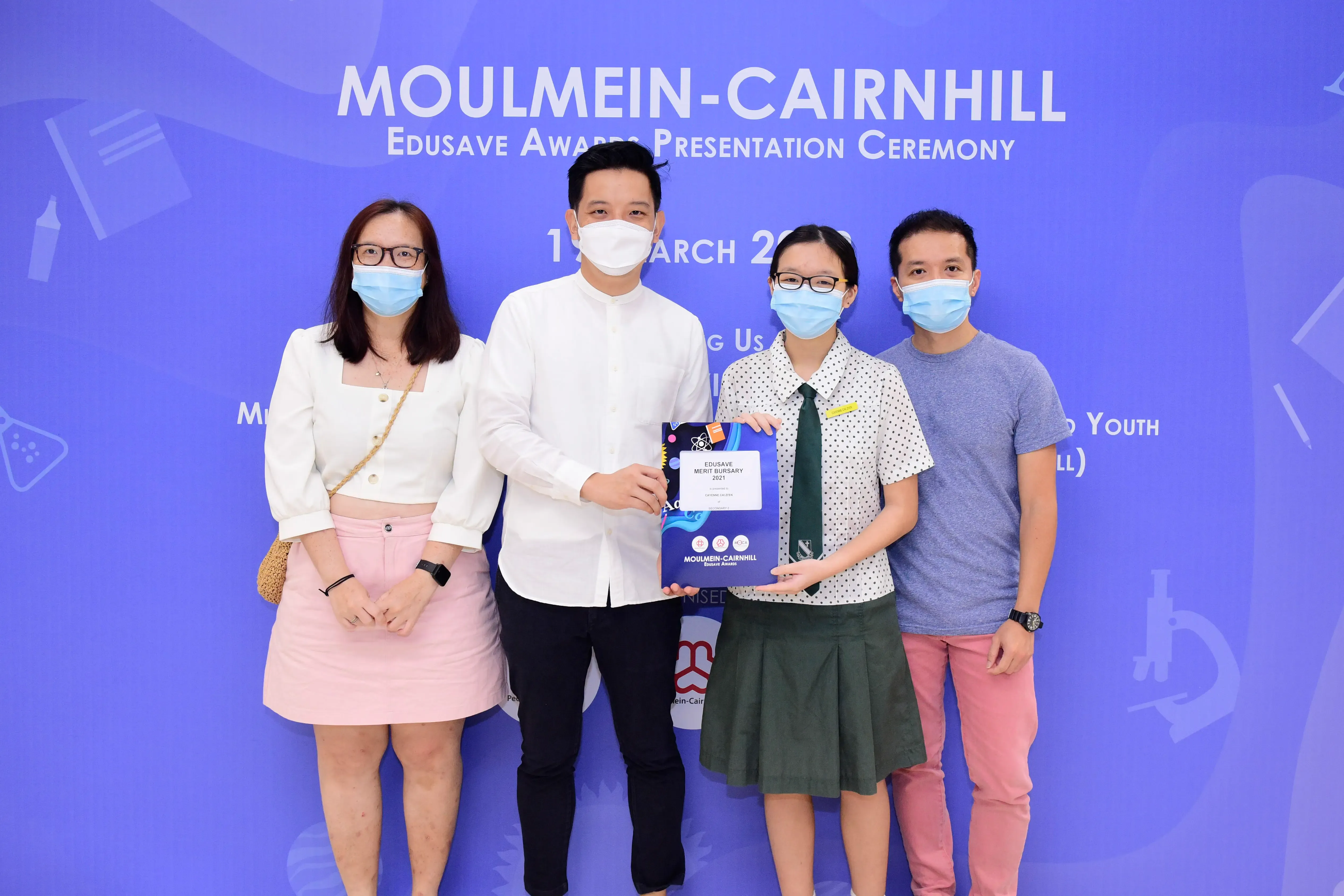 Four masked people pose at the Moulmein-Cairnhill Edusave Awards Presentation Ceremony, with a student holding an award.