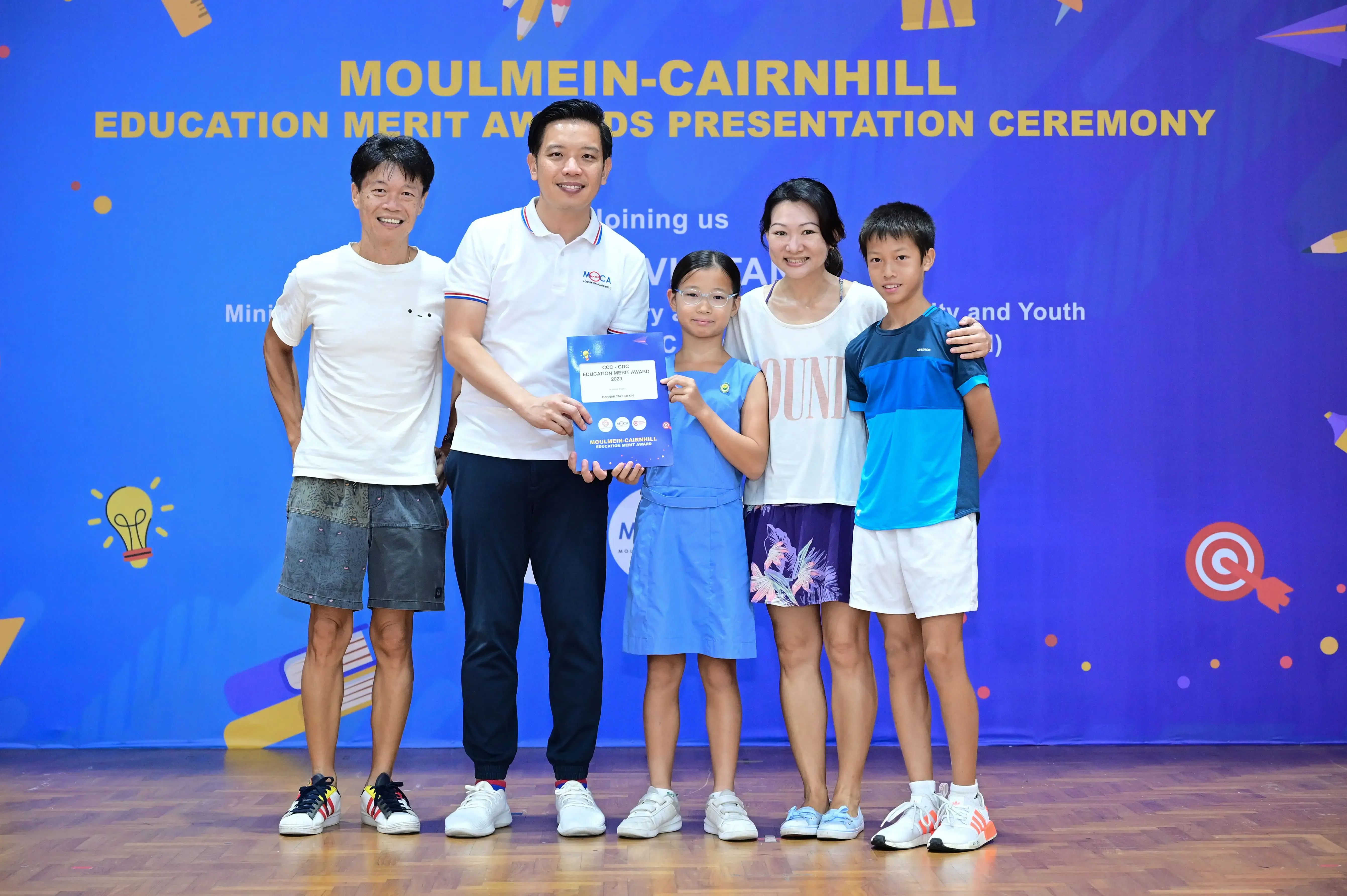 Five people pose at the Moulmein-Cairnhill Education Merit Awards Presentation Ceremony, with a girl holding an award certificate.